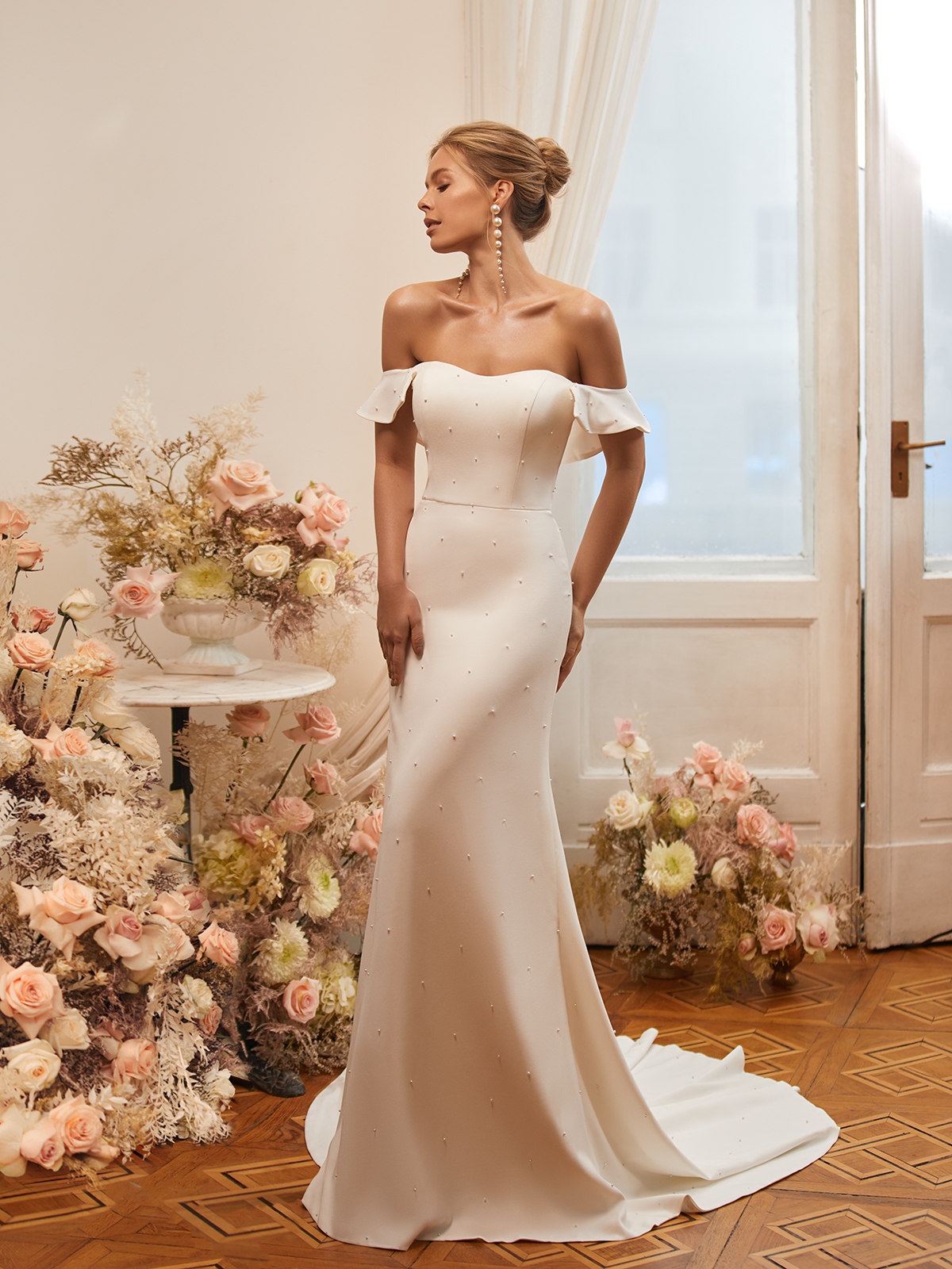Bride standing in front of flower arrangements in a romantic crepe wedding gown with pearls placed all over