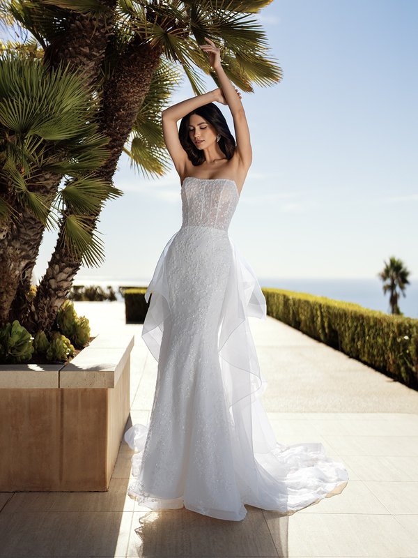 An elegant bride in Moonlight Couture H1585 a strapless wedding dress adorned with delicate floral embroidery and a flowing organza train, standing by the ocean 