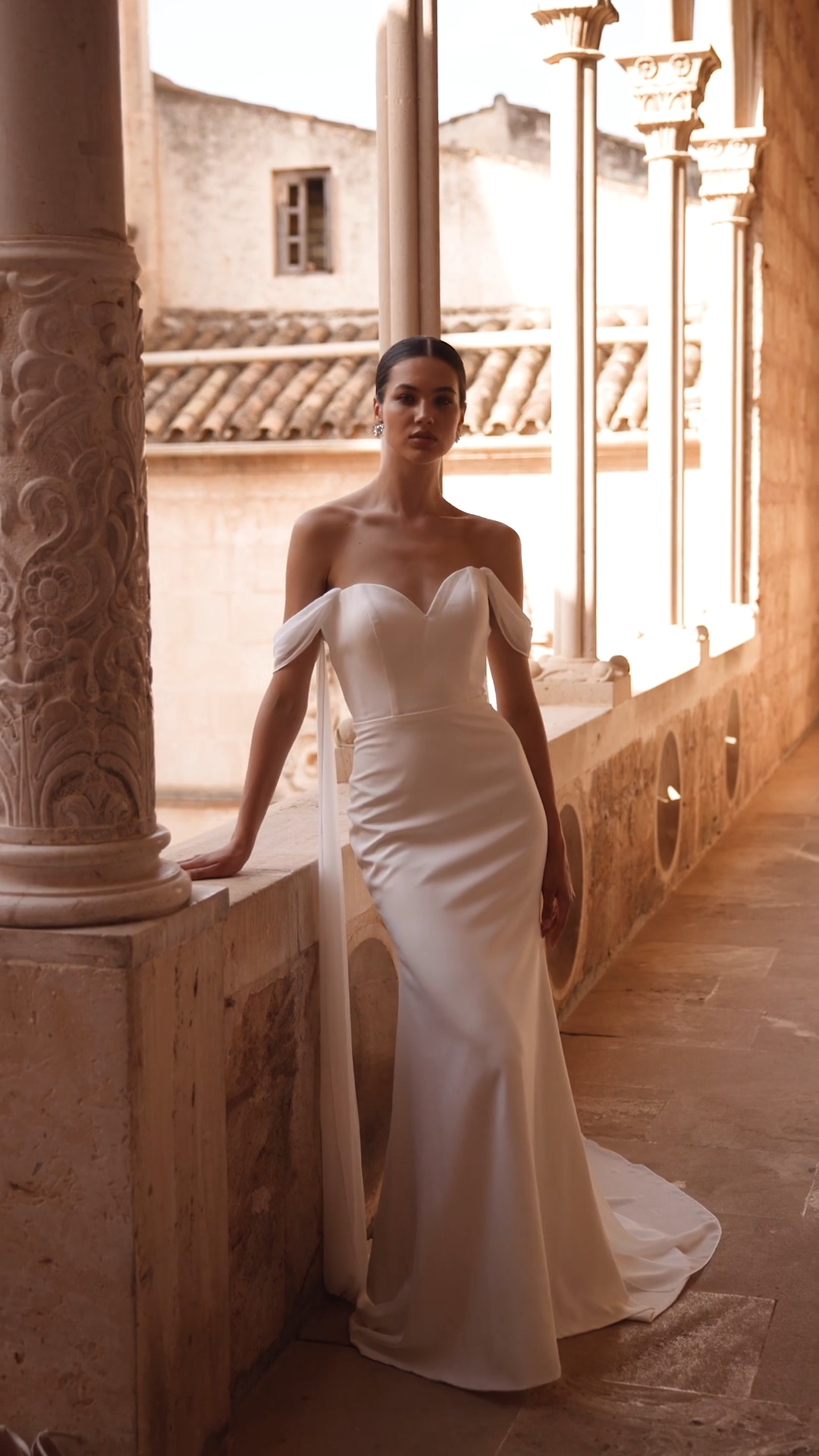 Bride leaning against wall in a ivory crepe mermaid wedding dress with chiffon off the shoulder sleeves