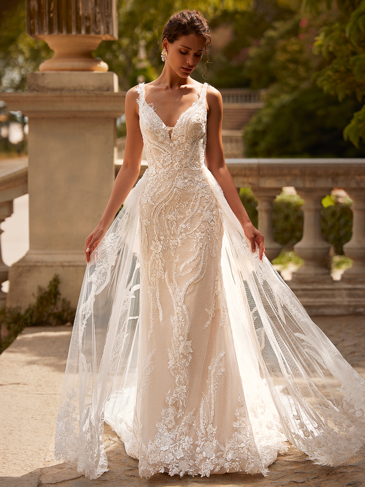 Woman standing in a sparkly lace wedding dress, holding onto its detachable train. 