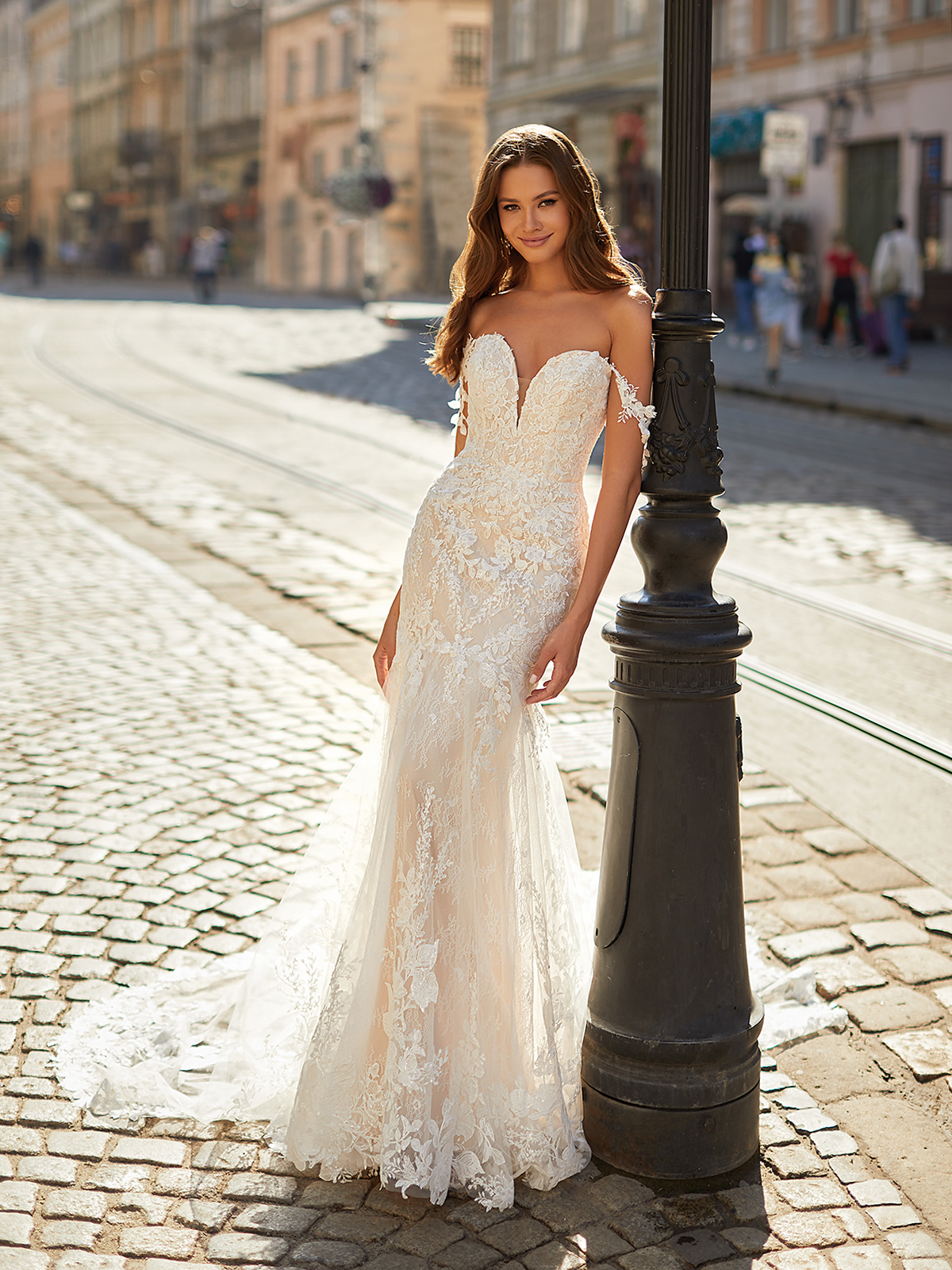 Bride leaning against an outdoor lamp post in a white wedding dress with off-the-shoulder swag sleeves