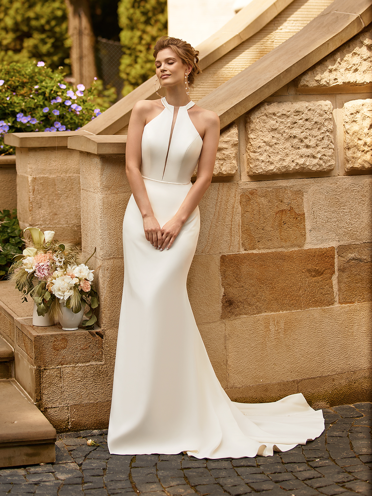Tall bride in a crepe wedding gown with a halter neckline standing next to outdoor staircase with flower arrangements