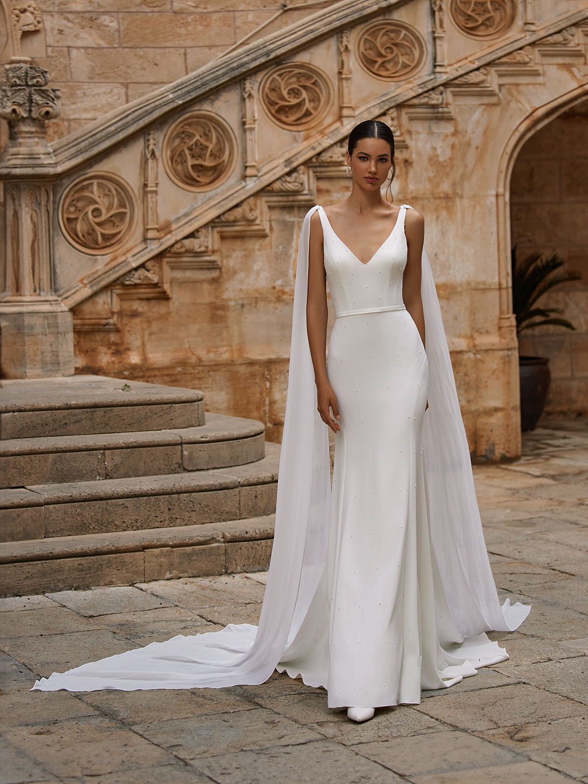 Bride standing in front of staircase in a romantic crepe wedding gown with pearls placed all over