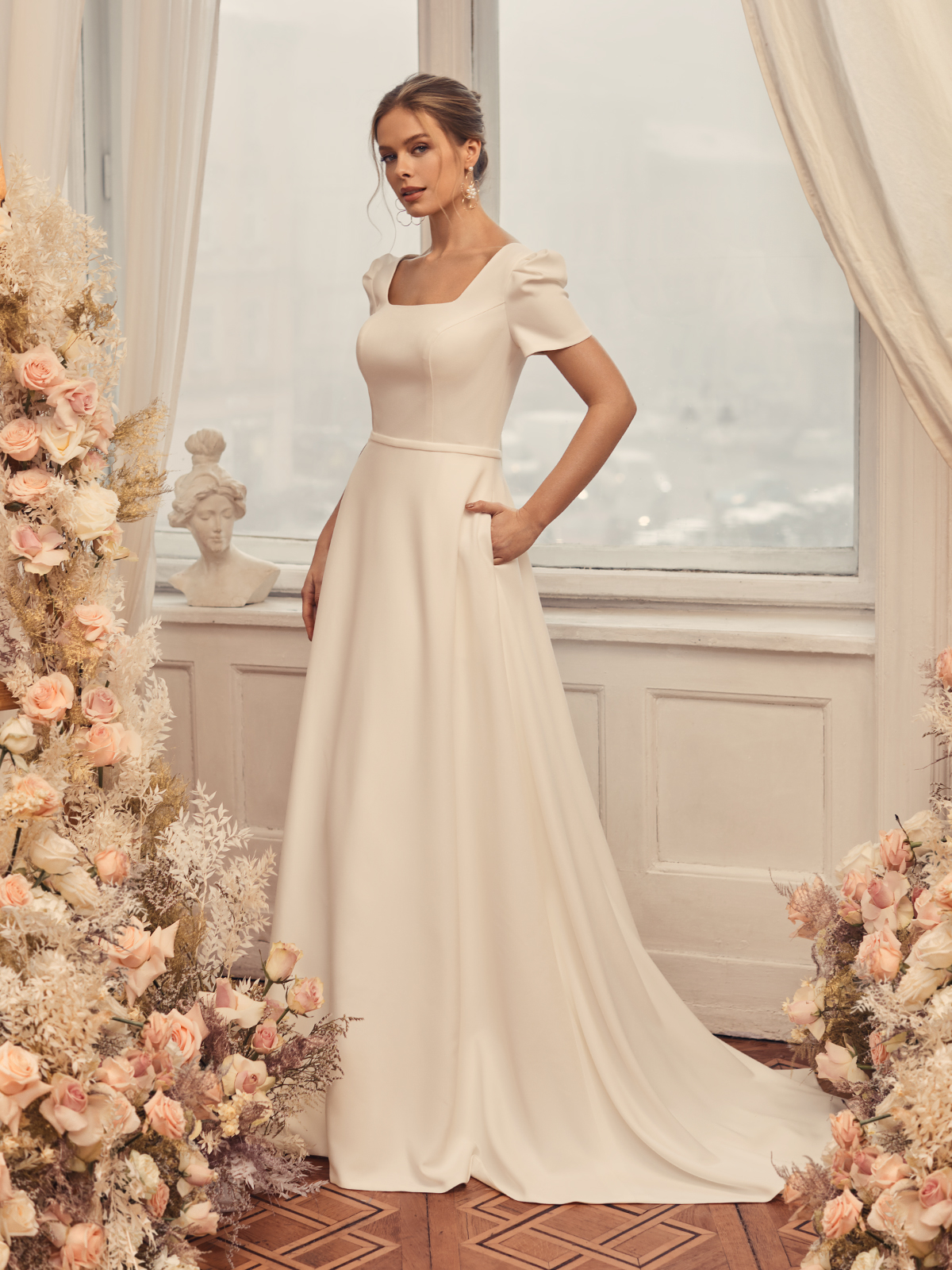 Woman standing next to flowers wearing an A-line bridal gown with a square neckline and short puff sleeves