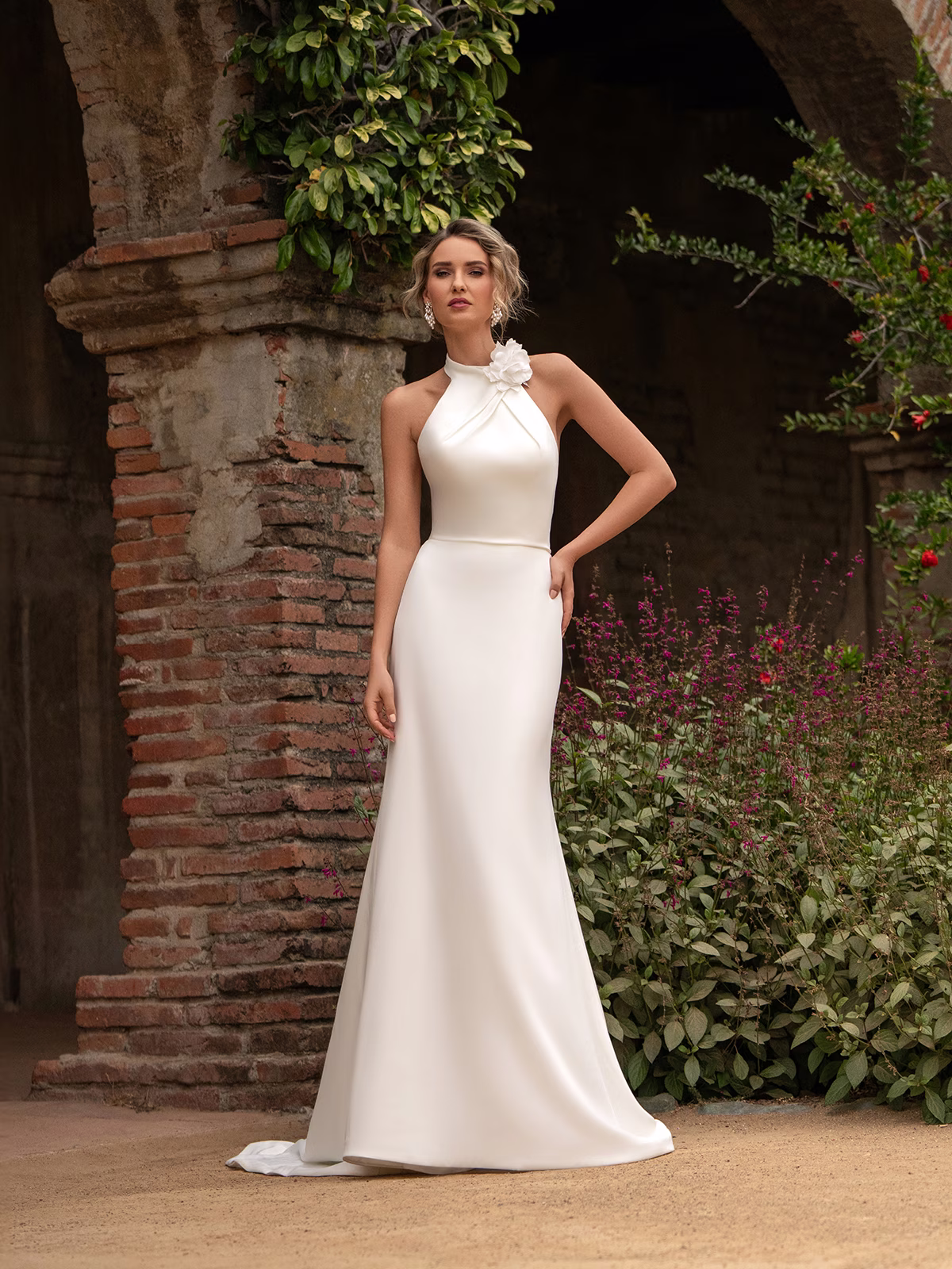 Tall bride in a crepe wedding gown with a halter neckline standing next to outdoor archways and foliage