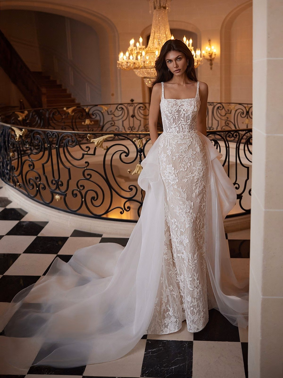 A bride in an elegant lace wedding gown with a long detachable organza train stands on a black-and-white checkered floor. A grand chandelier provides soft lighting Moonlight Couture H1651