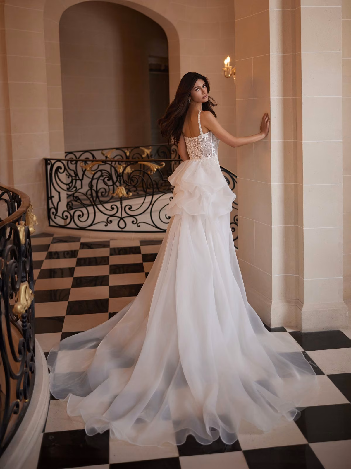 A bride in a flowing ivory gown stands on a black and white checkered floor of an elegant hallway. She touches the wall, exuding grace and elegance Moonlight Couture H1651