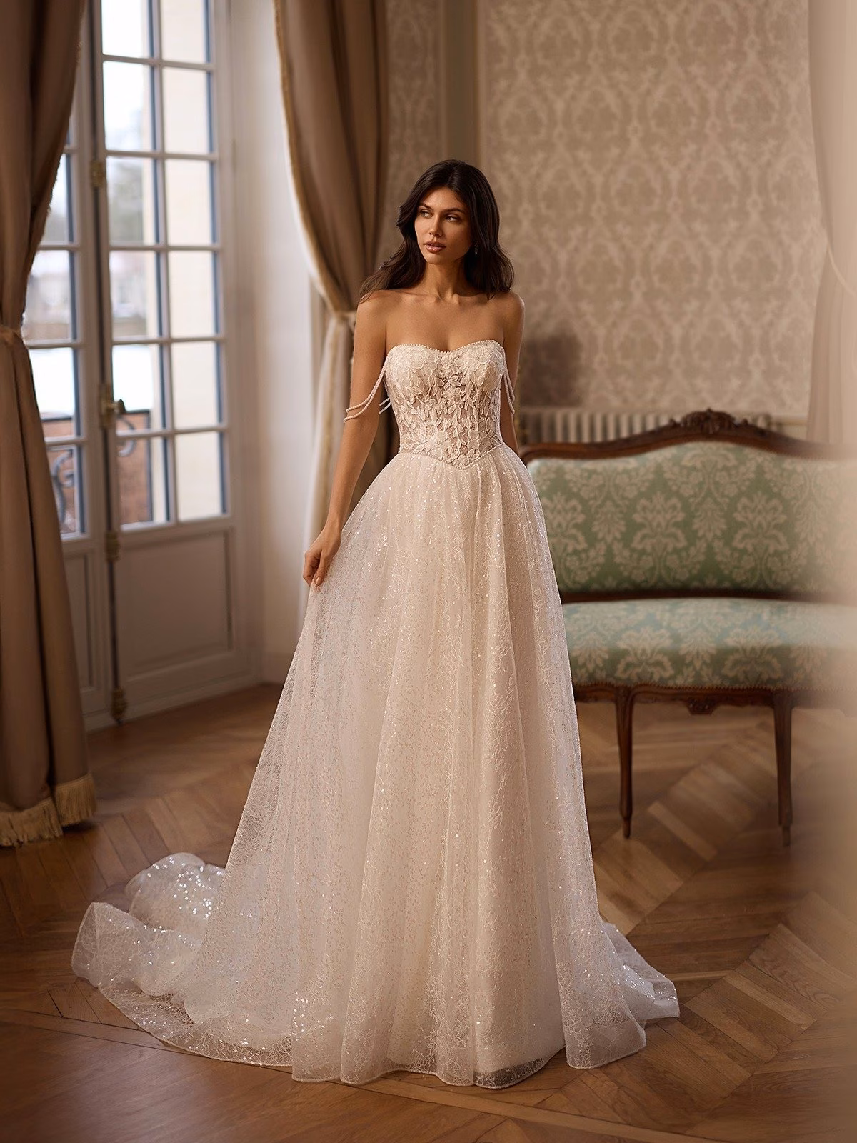 A bride in a sparkly Basque waist A-line and lace bridal gown with pearl strand sleeves stands in a classic room with ornate wallpaper and a vintage sofa Moonlight Couture H1652