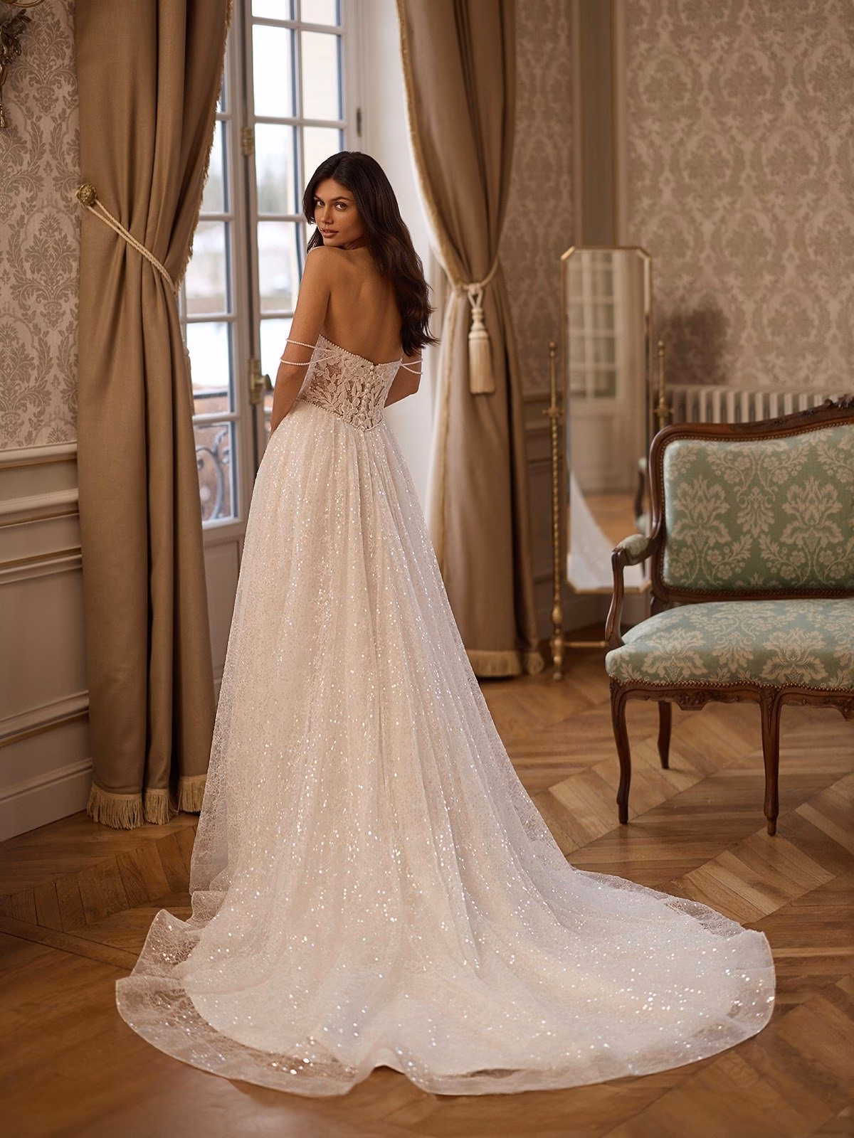 A bride in a lavish sparkling ivory wedding gown with open illusion back and pearl swag sleeves stands near a window in a vintage room with ornate wallpaper and draped curtain Moonlight Couture H1652