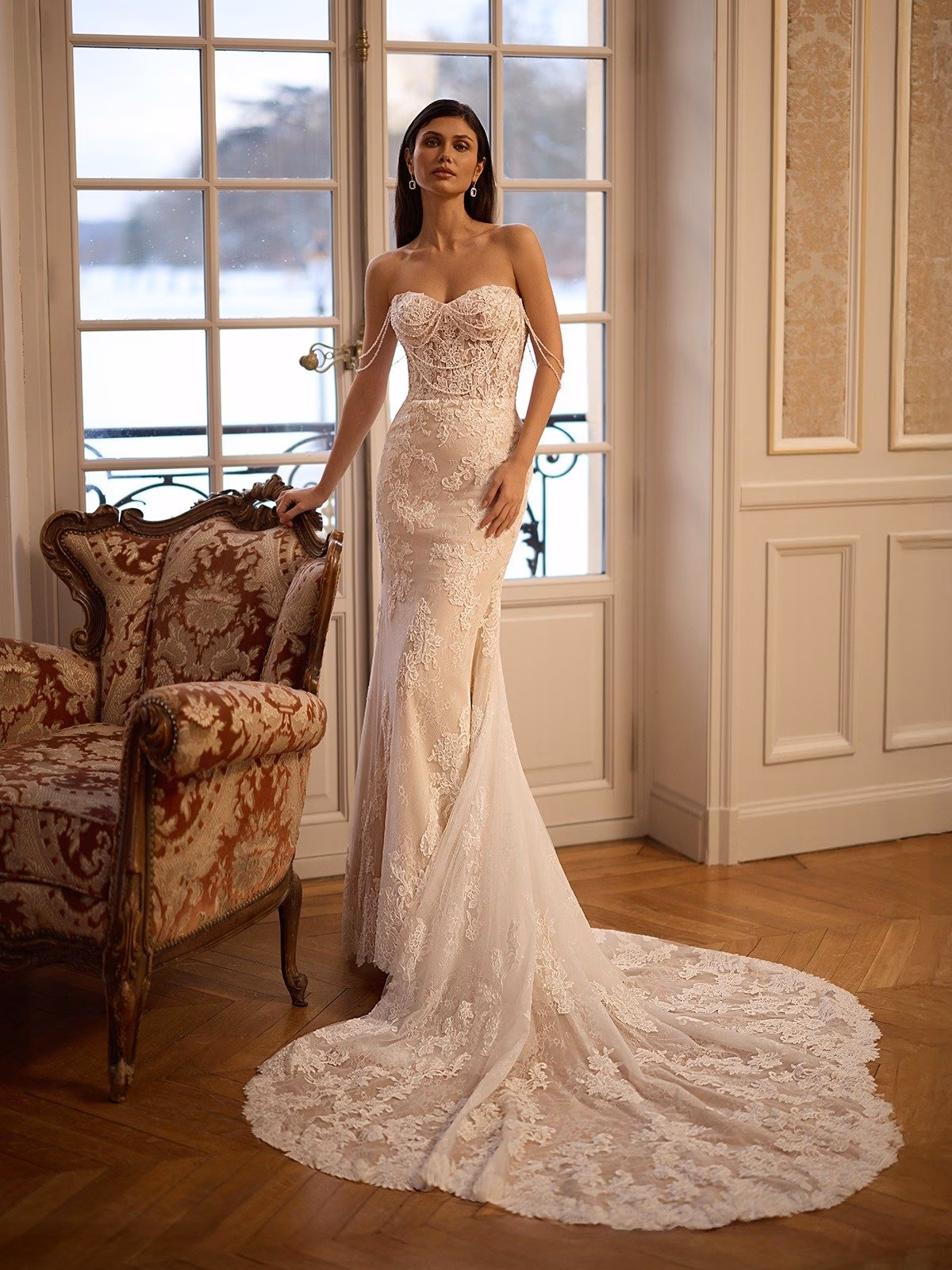 A bride in an elegant all lace wedding gown with beaded bodice stands by ornate French doors, next to an antique chair. The setting is sophisticated and serene Moonlight Couture H1653