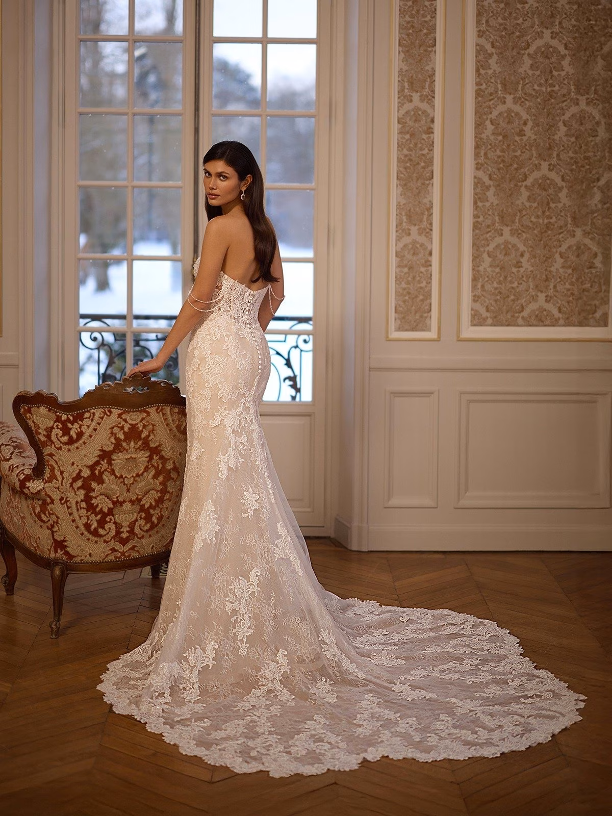 A bride in an elegant strapless illusion back lace wedding gown with long detailed lace train stands by a large window with a snowy view. The room features ornate wallpaper and an antique chair Moonlight Couture H1653