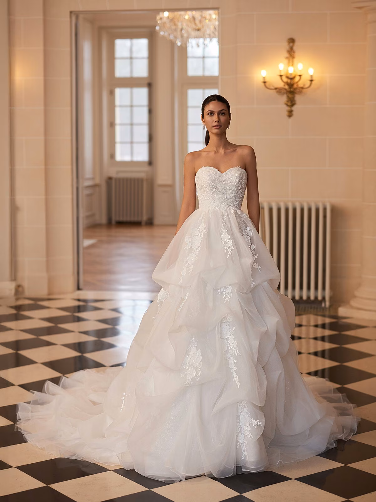 A bride stands in an elegant room wearing a strapless, lace-embellished ivory wedding gown with a voluminous pickup skirt. The floor is black-and-white checkered, and an ornate chandelier and wall sconce add a touch of opulence Moonlight Couture H1654