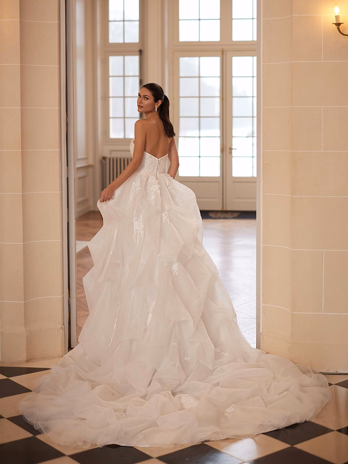 Bride in elegant strapless gown stands in grand hall, gazing back. Wedding dress has a flowing, ruffled train. Large windows and checkerboard floor create a classic, serene ambiance Moonlight Couture H1654