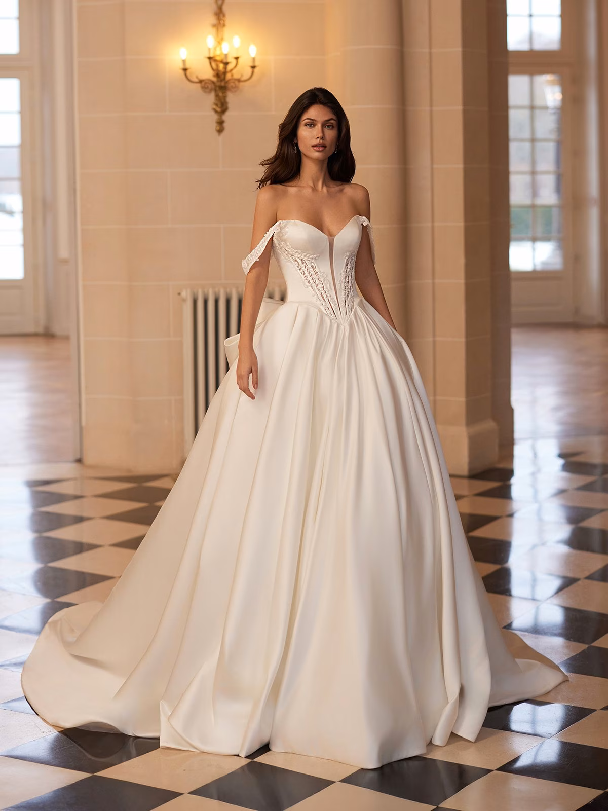 A bride stands elegantly in a spacious room with checkered flooring, wearing an off-shoulder ball gown with lace details. The mood is sophisticated and serene Moonlight Couture H1655