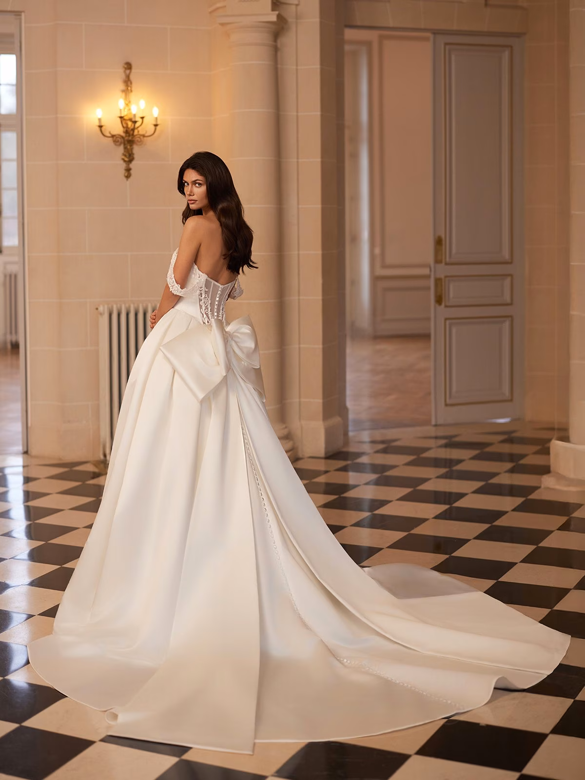 A woman in an elegant satin wedding gown with a long train and large bow stands in a grand room with checkered floor. She gazes back, exuding grace Moonlight Couture H1655