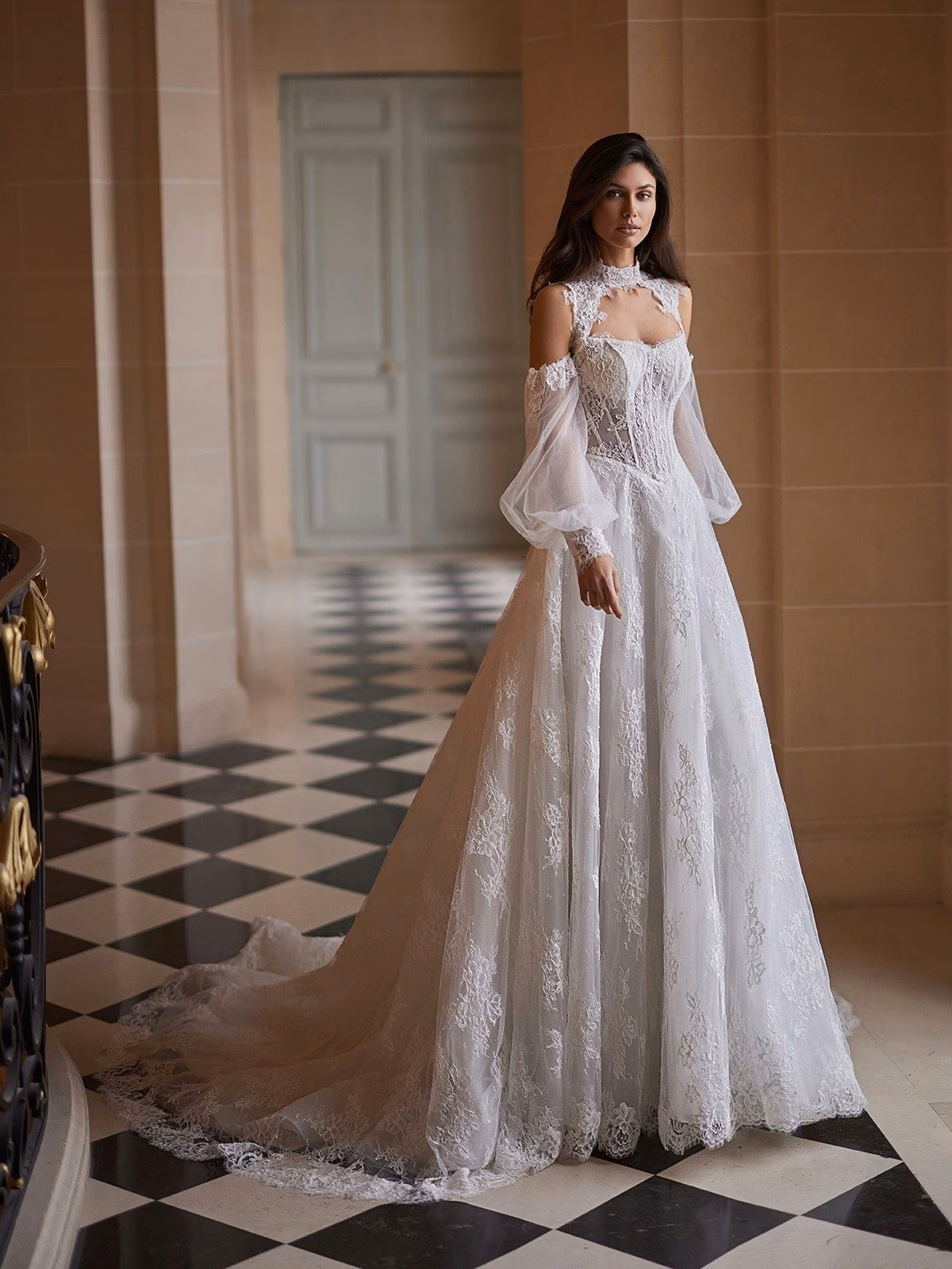 A bride stands in an elegant setting wearing an all lace A-line wedding gown featuring a lace and tulle bolero jacket and a flowing train. The ambiance is sophisticated and serene Moonlight Couture H1657