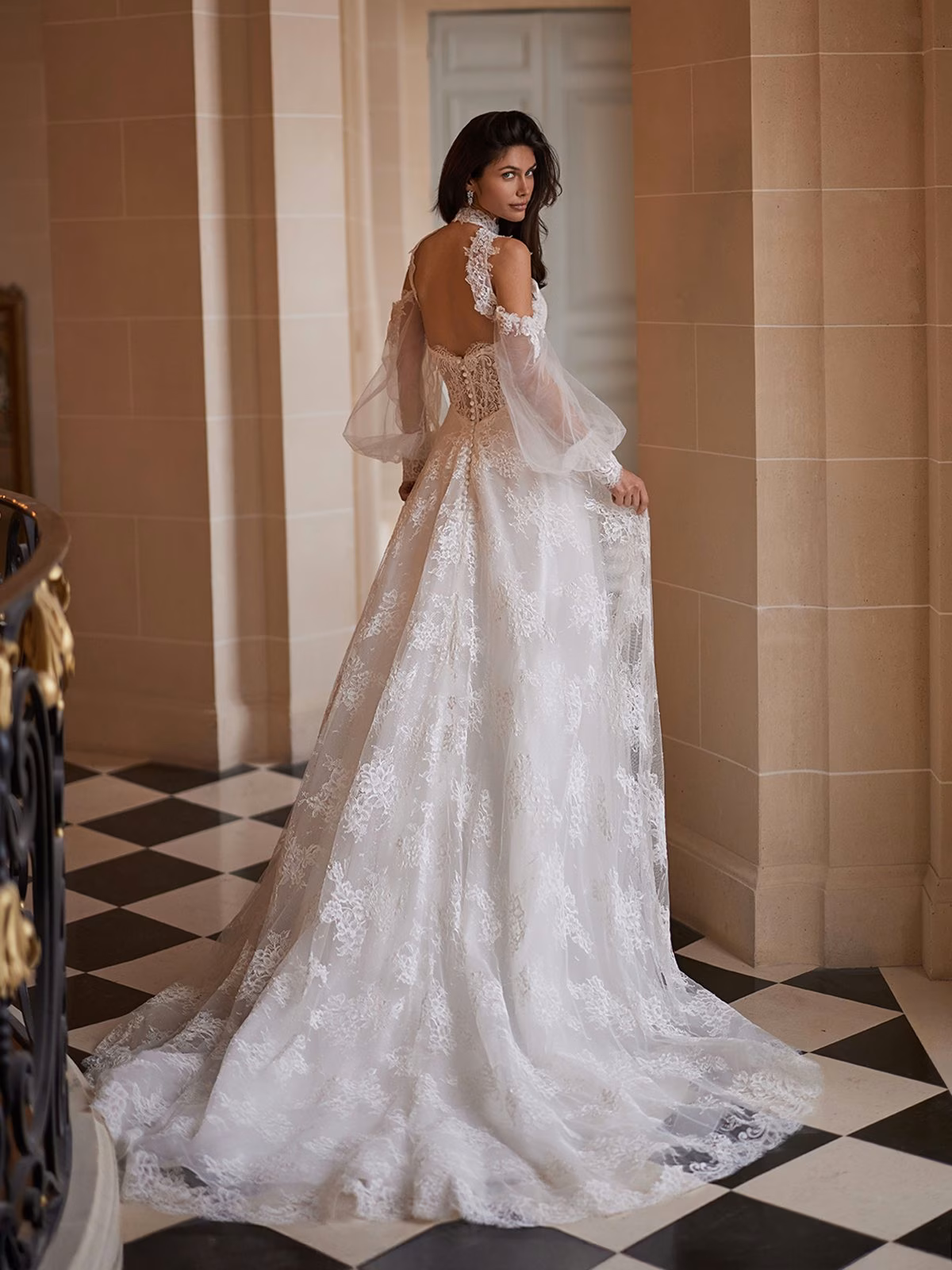 A bride in an elegant lace wedding dress with open back stands gracefully in a grand, marble-tiled hallway, exuding a sense of timeless elegance and romance Moonlight Couture H1657