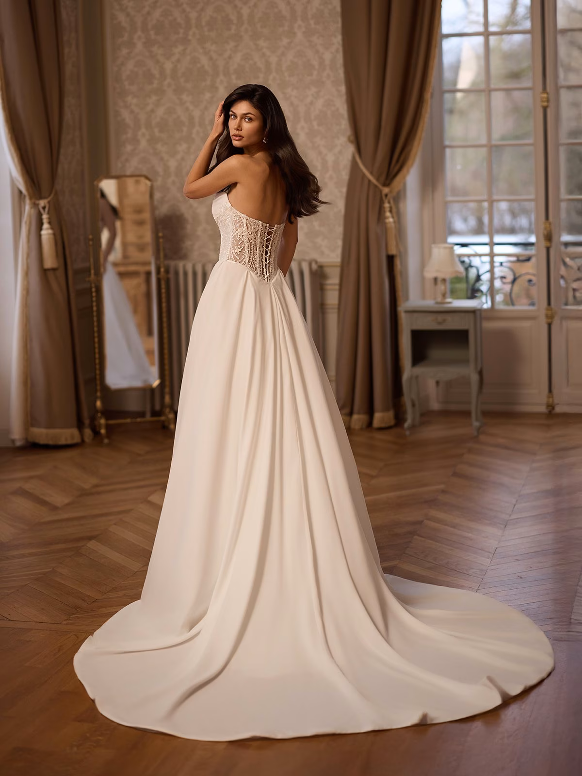 Elegant bride in an ivory strapless wedding gown with lace corset back, standing in a vintage room with wooden floor, curtains, and tall windows Moonlight Couture H1658