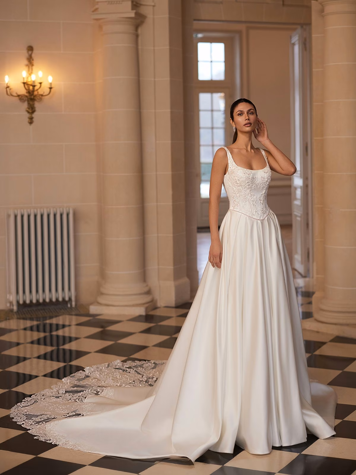 A bride in a scoop neck with straps, satin A-line wedding gown with a long train and lace details stands in a vintage room Moonlight Couture H1659