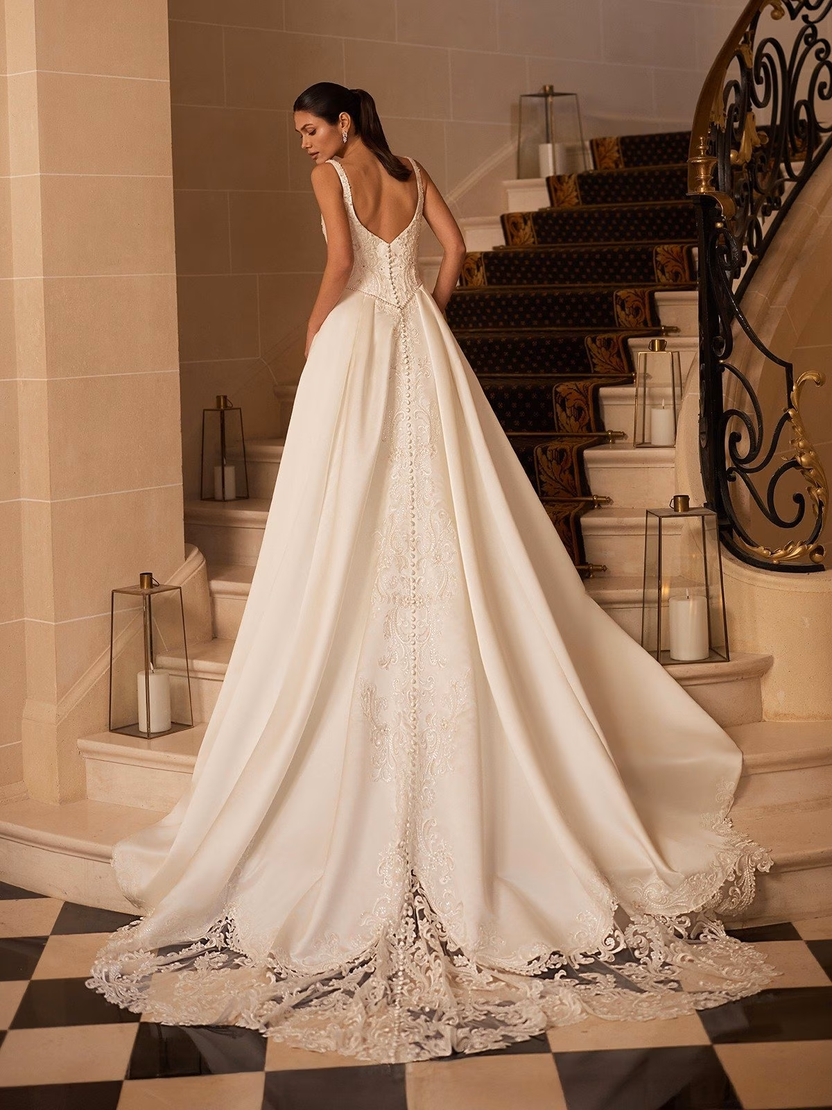 A bride in a flowing satin A-line wedding gown with long lace detailed train stands on a checkered floor beside a grand staircase adorned with ornate lanterns, exuding elegance Moonlight Couture H1659