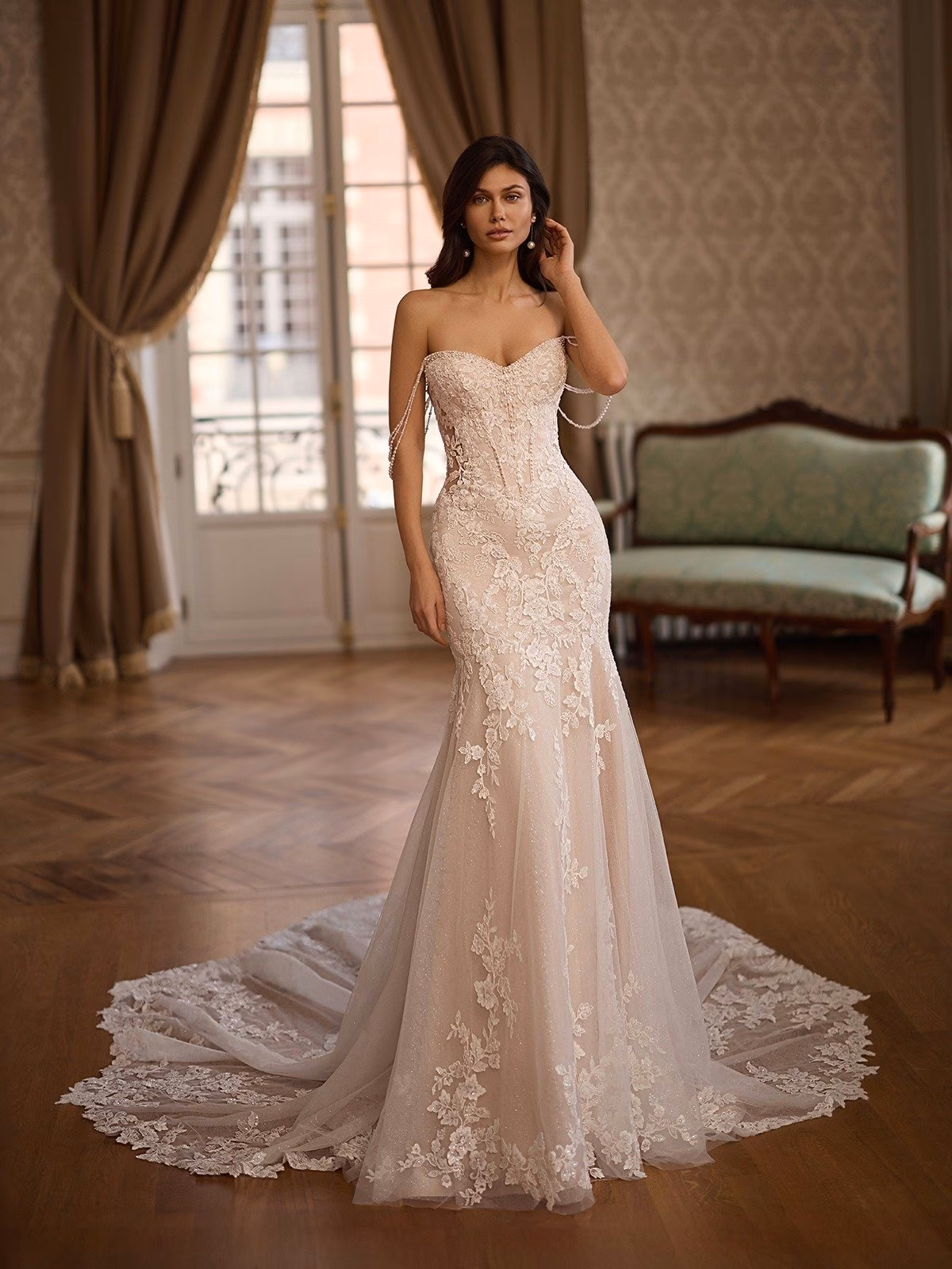 A bride stands in an ornate room wearing a deep sweetheart with pearl beaded swag sleeves, lace wedding gown with a long train. The setting adds a vintage, sophisticated feel Moonlight Couture H1666