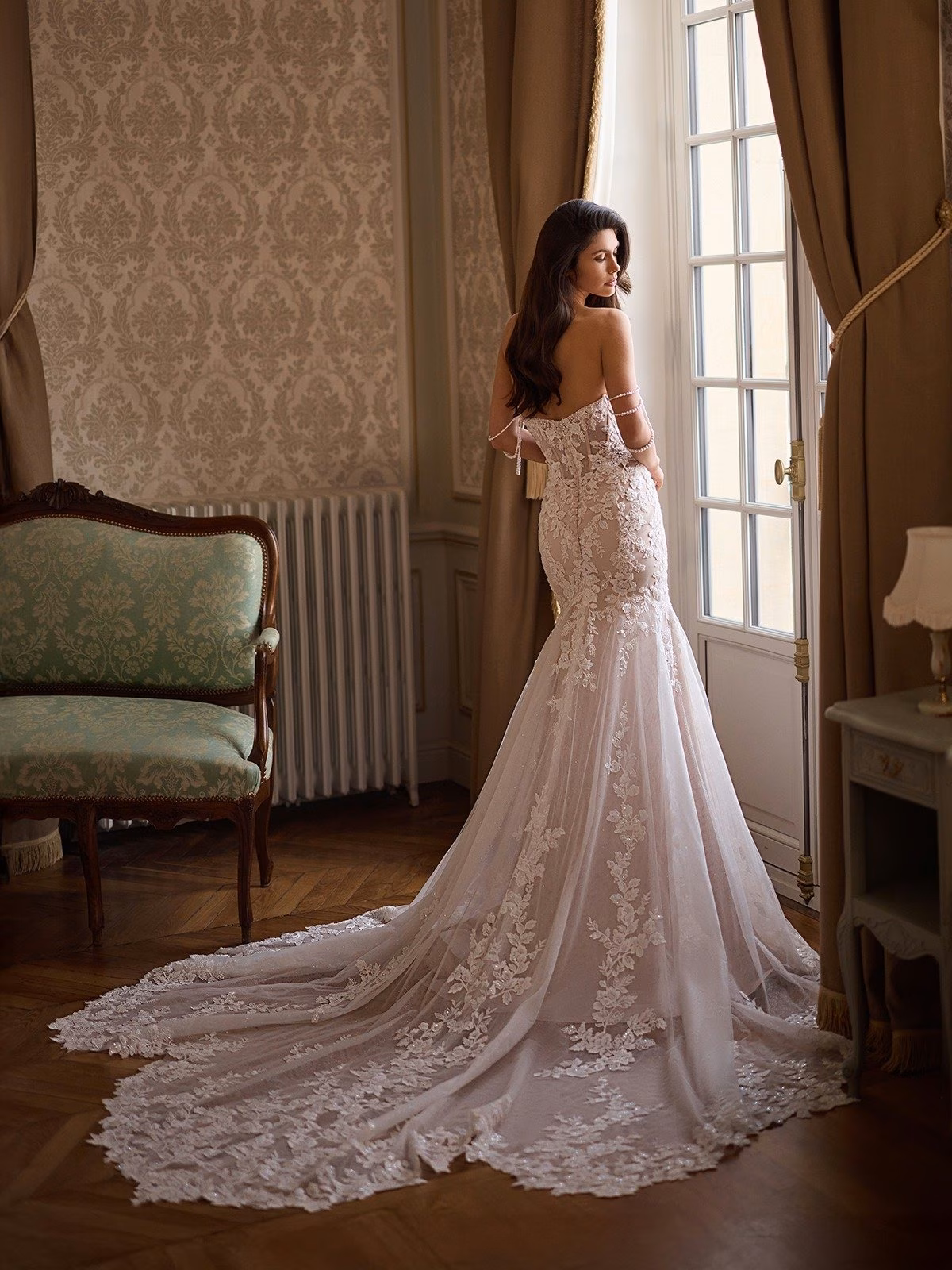 A bride in an elegant lace wedding gown with long lace detailed train stands gazing out a tall window. The room