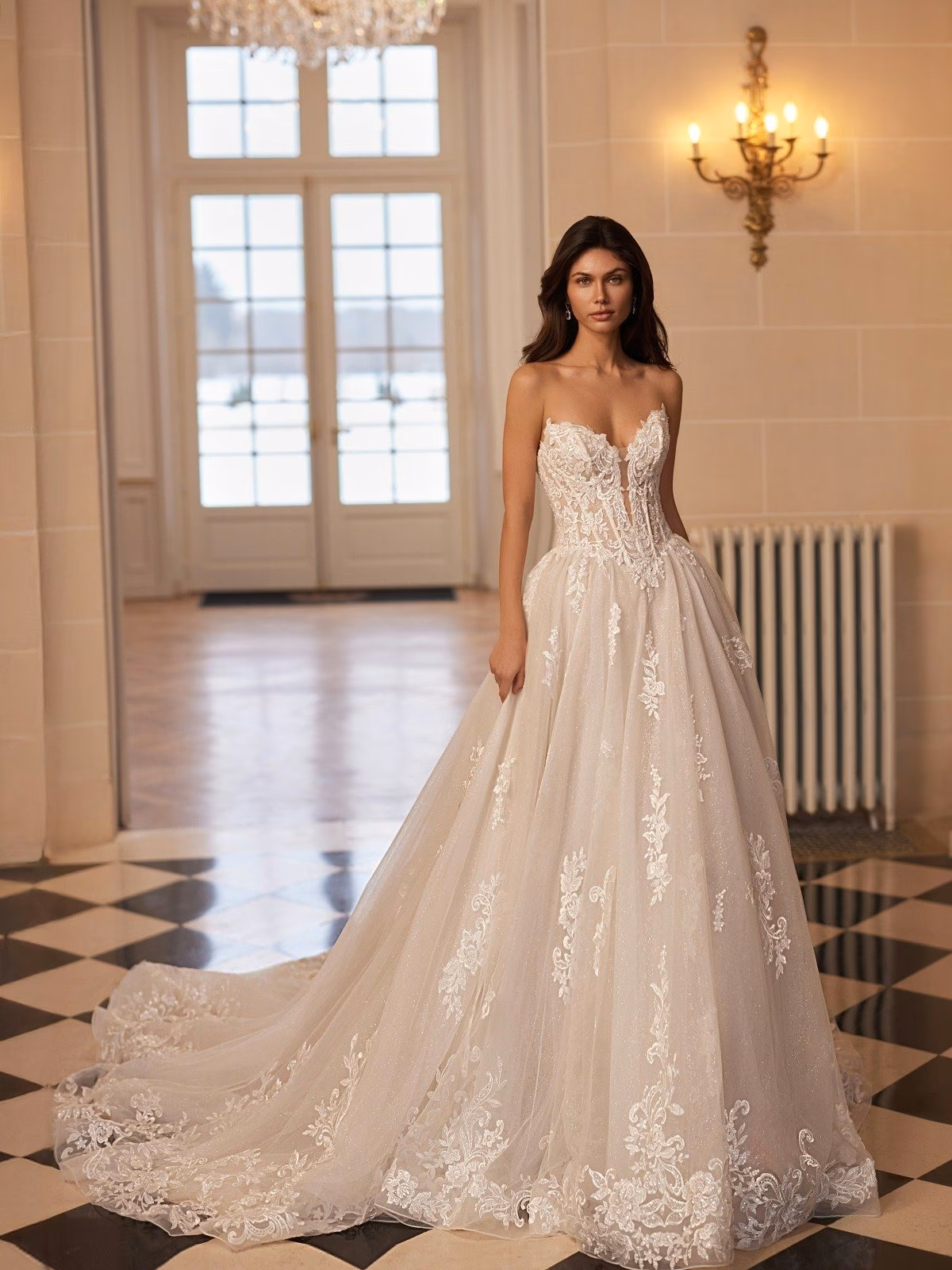 A bride wearing a lavish lace wedding gown with a fitted deep sweetheart bodice and flowing tulle ball gown skirt stands on a checkerboard floor. Elegant chandeliers and soft lighting create a romantic ambiance Moonlight Couture H1661