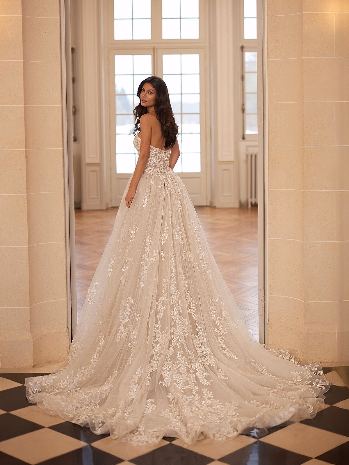 A bride in an elegant lace ball gown wedding gown stands in a grand room. The dress features intricate lace and a long lace train. The setting has a luxurious, serene ambiance Moonlight Couture H1661