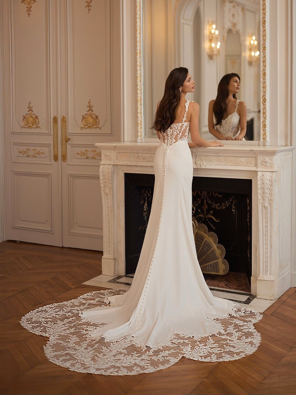 A bride in a crepe wedding dress with lace detailed train stands before a vintage mirror above a fireplace. The room features ornate decor and soft lighting Moonlight Collection J7002