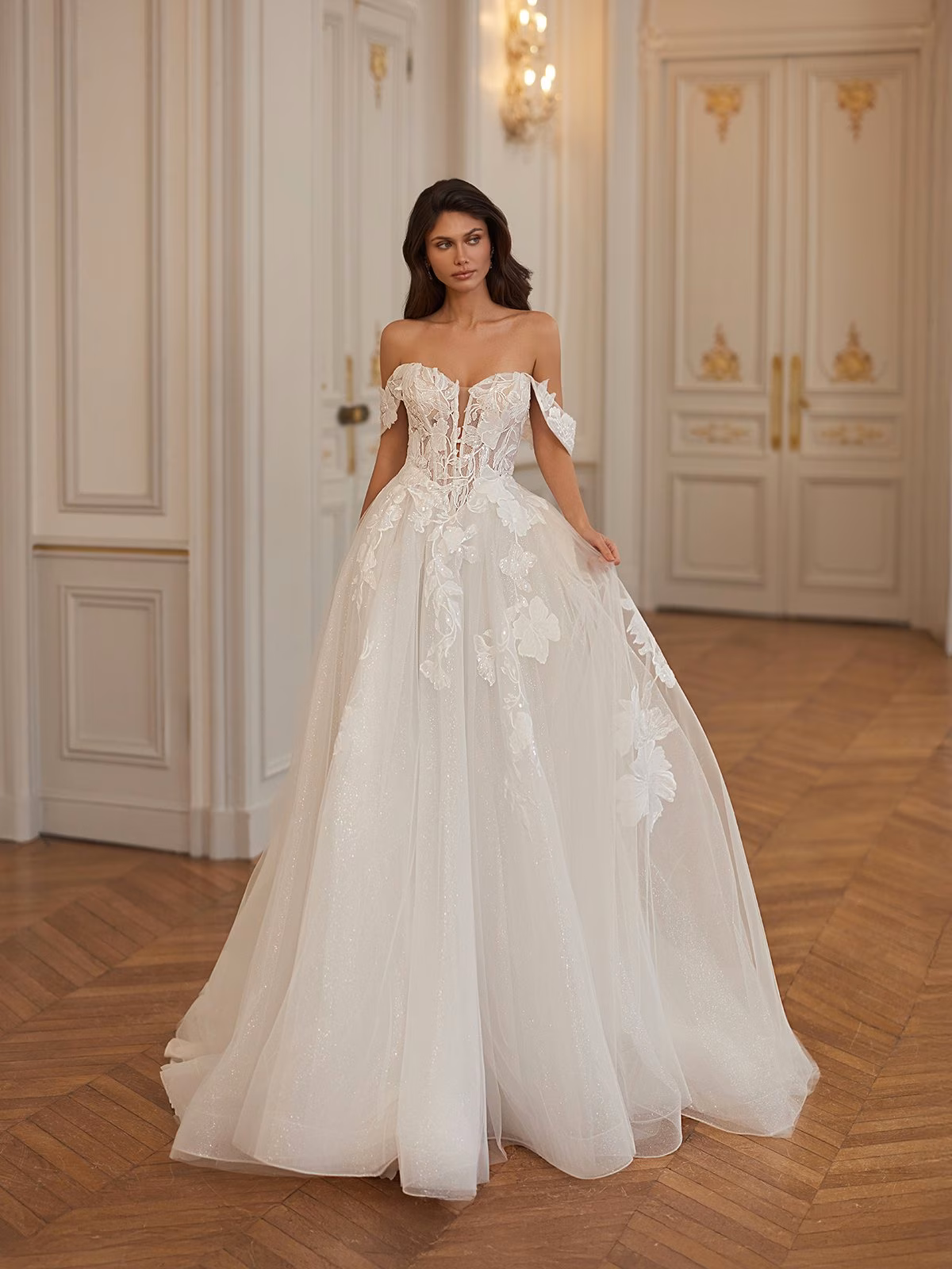 A bride walks in an elegant, off-the-shoulder, ivory tulle wedding gown with floral appliques and Basque waist on a wooden floor in a luxurious room with ornate doors and lighting Moonlight Collection J7003