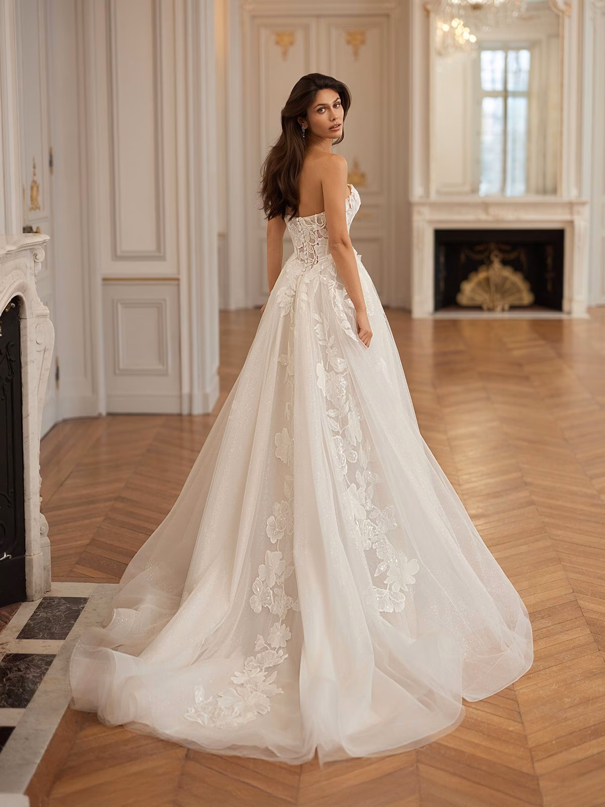 Bride in elegant strapless tulle wedding gown with lace details, stands in luxurious room with wood floors, chandeliers, and ornate fireplace, exuding a serene elegance Moonlight Collection J7003