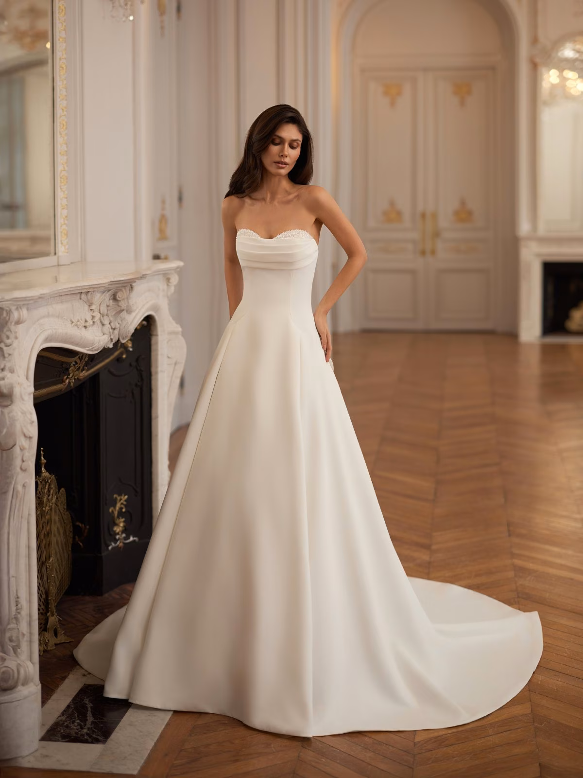 A bride stands elegantly in a luxurious, ornate room, wearing a strapless ivory double face crepe wedding gown with a flowing train and beaded details along the neckline. The background features a vintage-style fireplace and decorative doors, highlighting a sense of opulence Moonlight Collection J7004