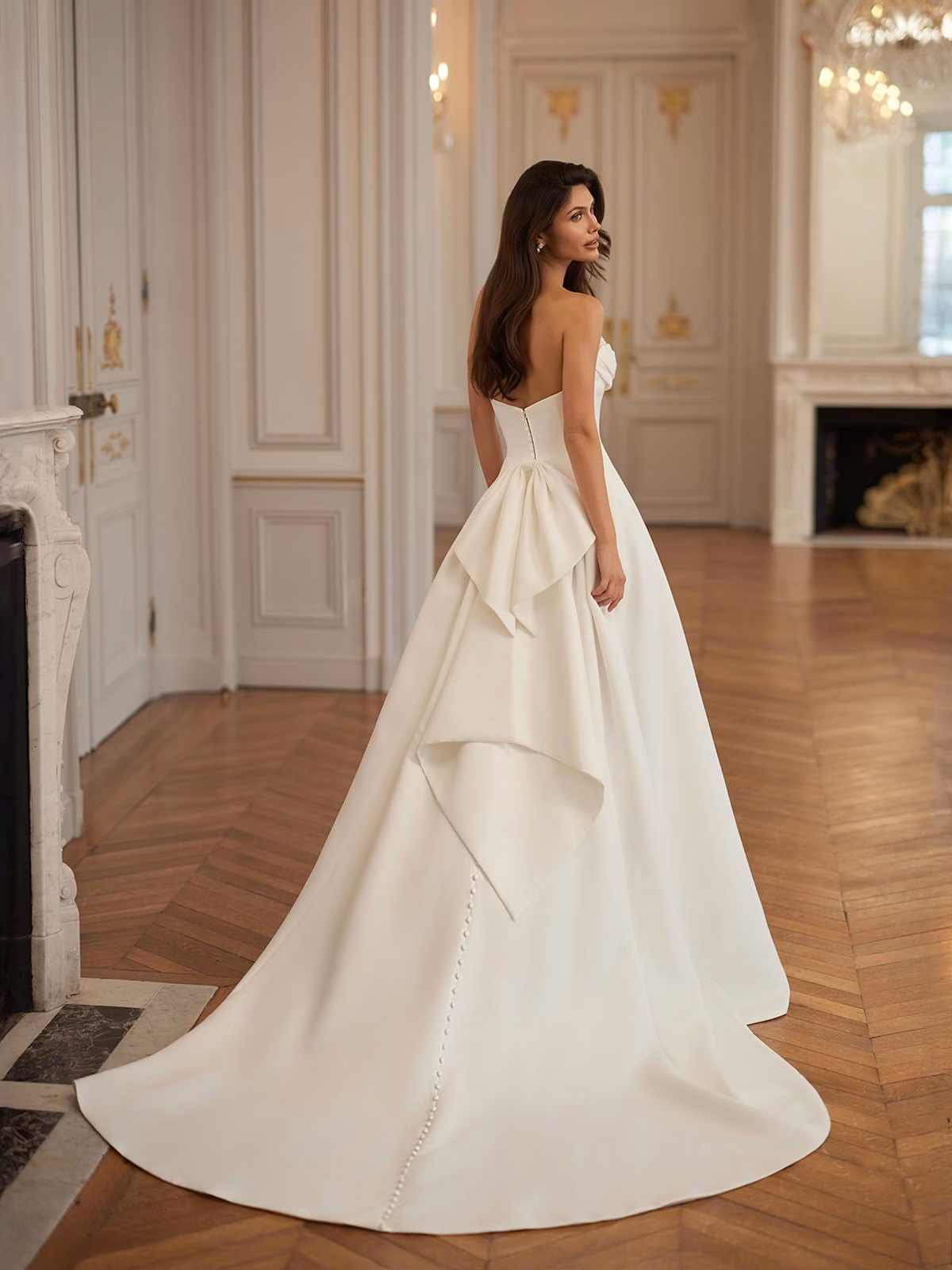 A woman in an elegant, strapless double face crepe wedding gown with a long train and statement bow stands in a luxurious room with ornate doors and hardwood floors, exuding sophistication Moonlight Collection J7004