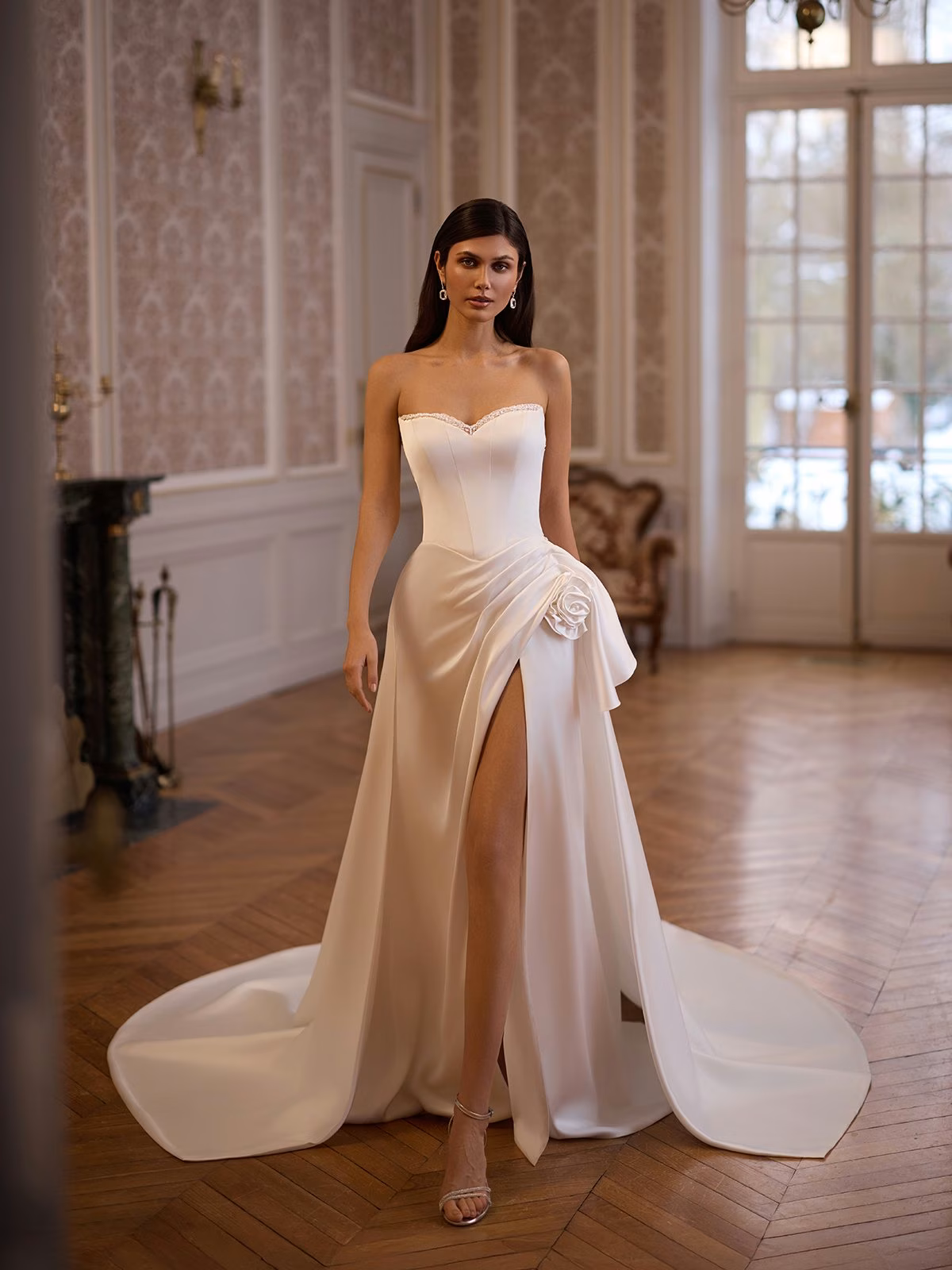 Bride in an elegant stretch satin strapless wedding dress with a high slit and floral accent, standing in a luxurious room with ornate walls and wooden floor Moonlight Collection J7005