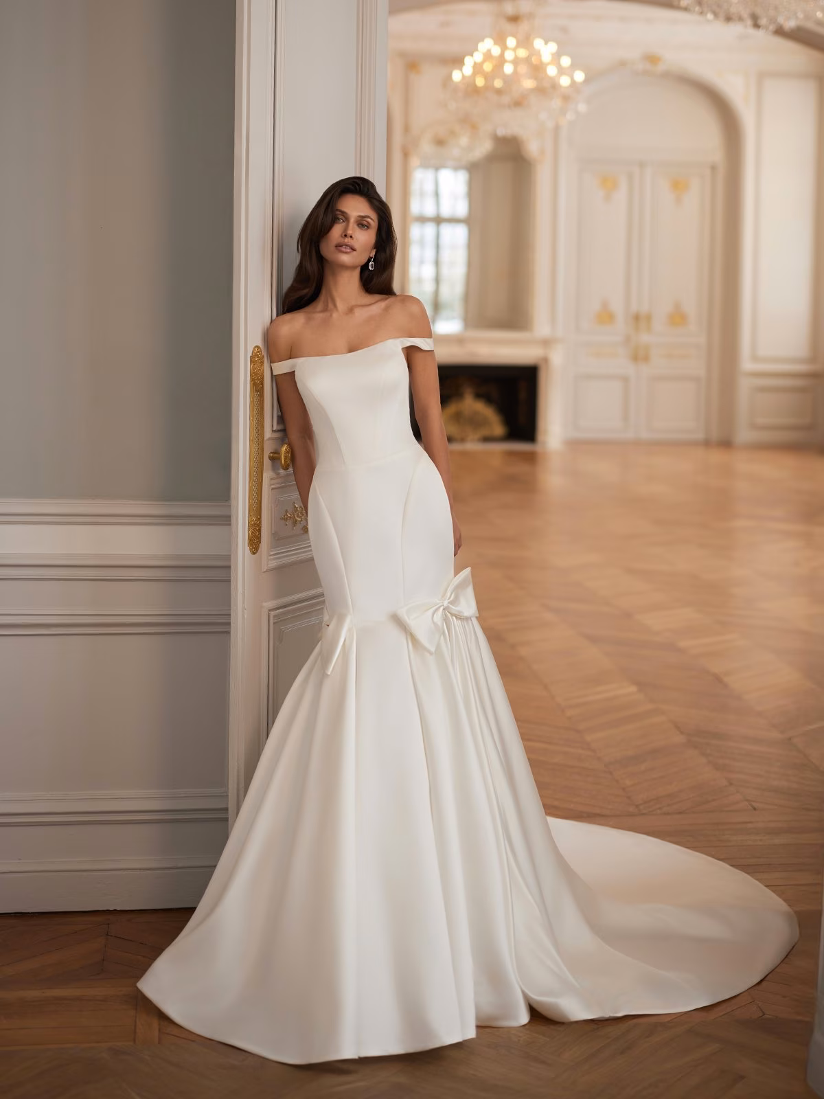 Bridal model in an elegant off-the-shoulder satin wedding gown with a mermaid silhouette and bow, standing in a luxurious room with chandeliers and parquet floors Moonlight Collection J7006
