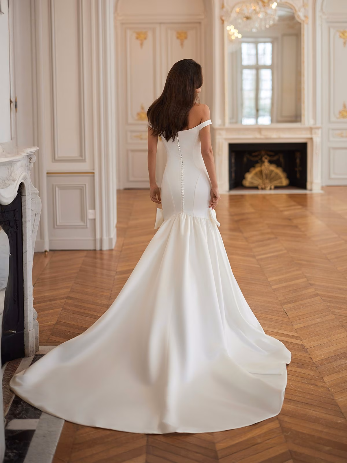 A bride in an off-the-shoulder, elegant satin mermaid wedding dress stands in a spacious, ornate room with parquet flooring. The dress flows into a long train, exuding grace and sophistication Moonlight Collection J7006
