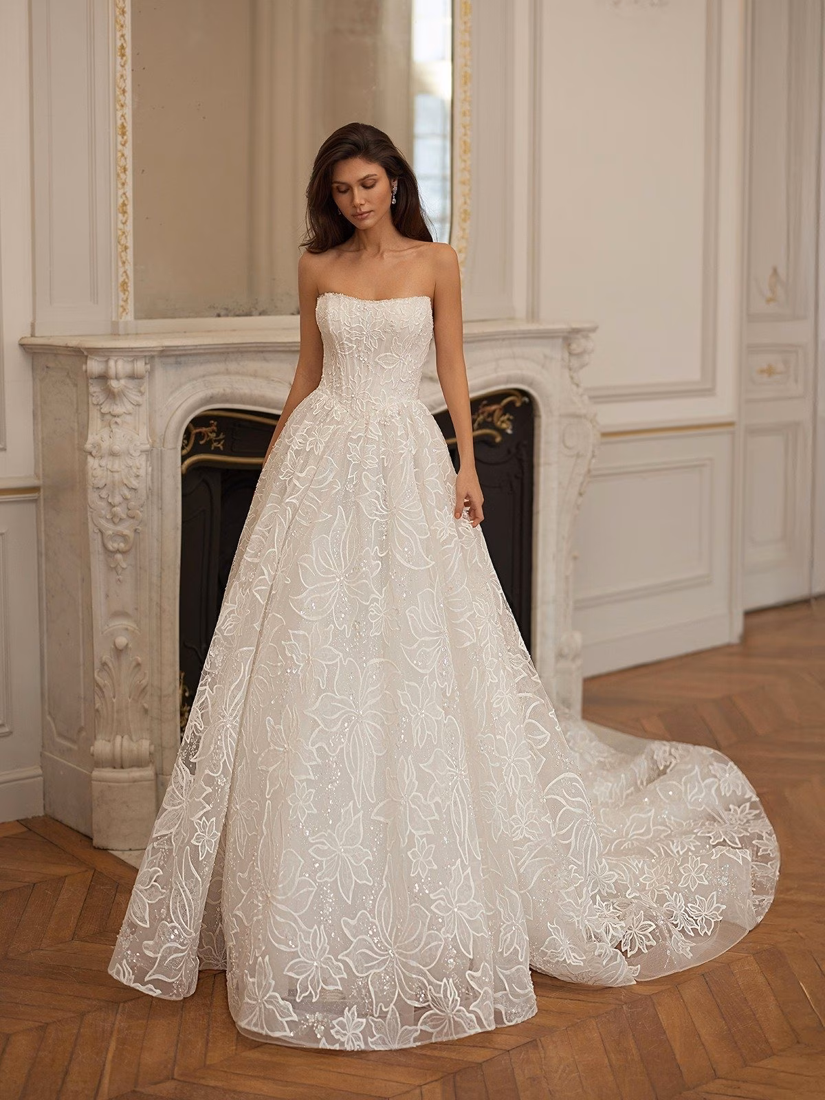 A bride in a beaded scoop neckline and floral lace A-line wedding gown with a corset bodice in a classic room with a marble fireplace, exuding grace and sophistication Moonlight Collection J7007