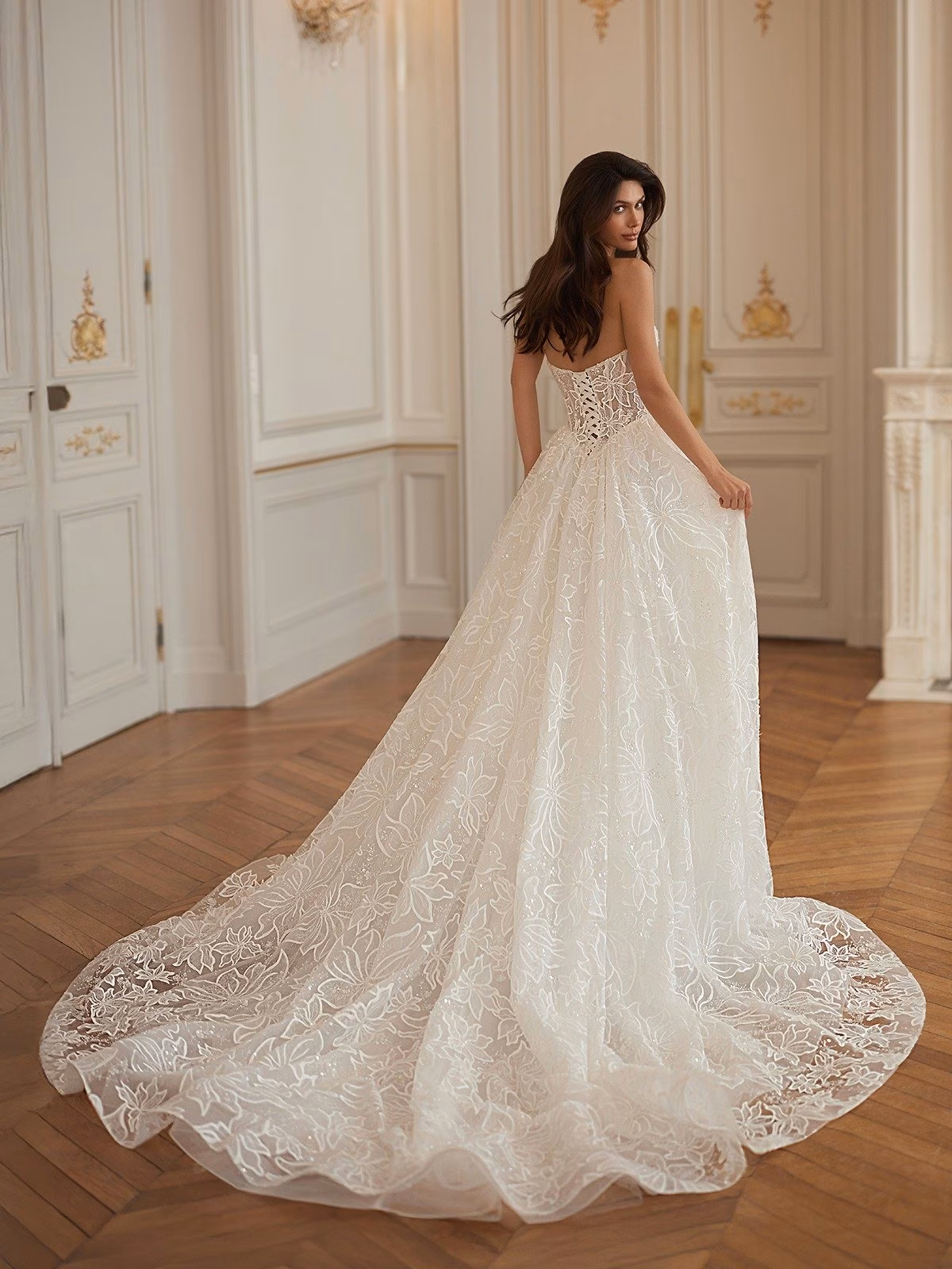 Bride in a lavish room, wearing an elegant strapless sparkly floral lace wedding gown with a long floral train. The setting feels classic and sophisticated Moonlight Collection J7007