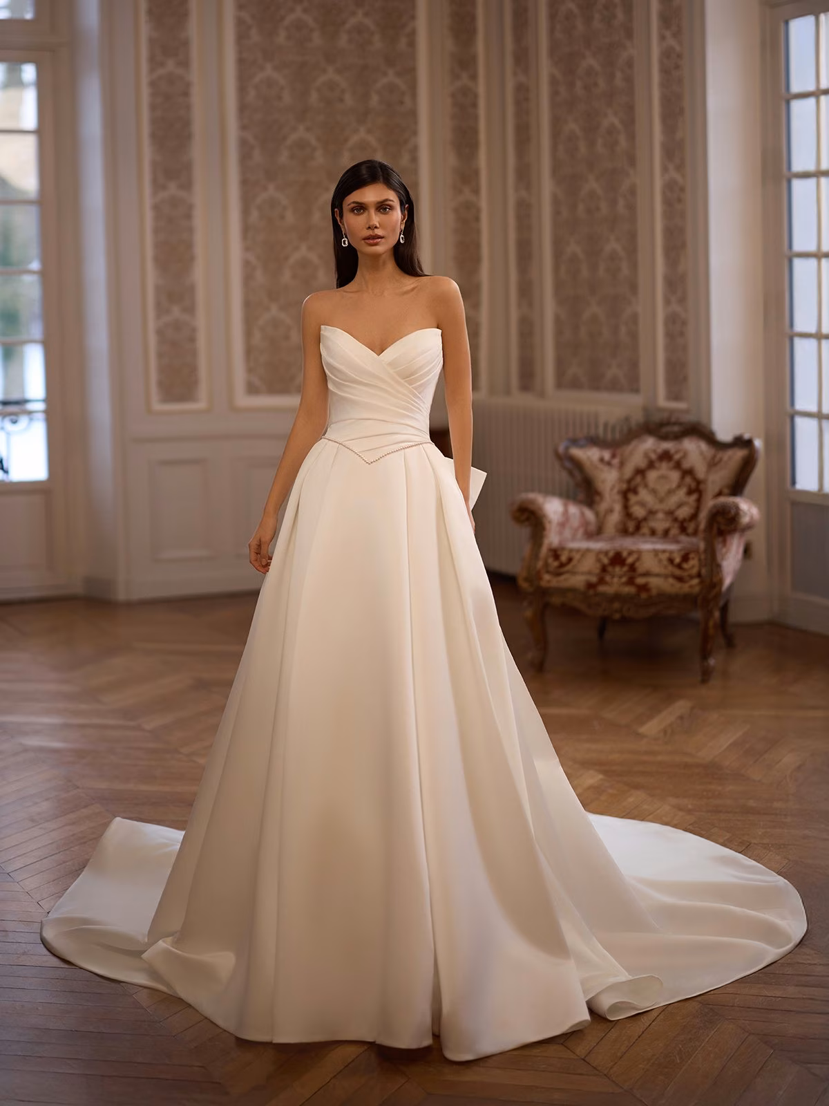 Bride in elegant pointed sweetheart ivory satin wedding gown with Basque waist stands in a lavishly decorated room with wooden floors and ornate wallpaper, exuding grace and sophistication Moonlight Collection J7008