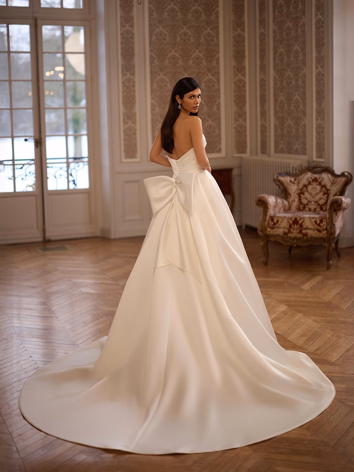 A bride in a strapless ivory satin A-line wedding gown with a large bow at the back stands in an elegant room. Light filters through tall windows, creating a serene ambiance Moonlight Collection J7008