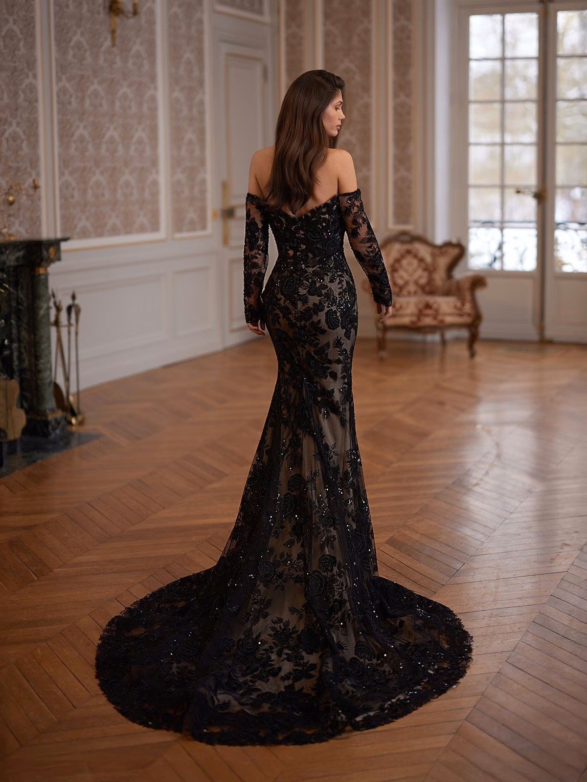 Bride in a black lace mermaid wedding gown with long sleeves stands in an ornate room with wooden floors and intricate wallpaper. The atmosphere is luxurious and sophisticated Moonlight Collection J7009