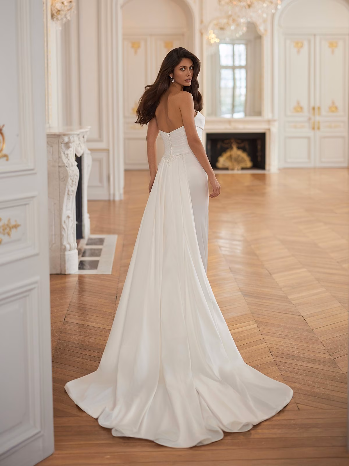 A bride in a strapless stretch satin ivory wedding gown with a long train stands in an elegant room with wooden floors, chandeliers, and large windows, exuding elegance Moonlight Collection J7010