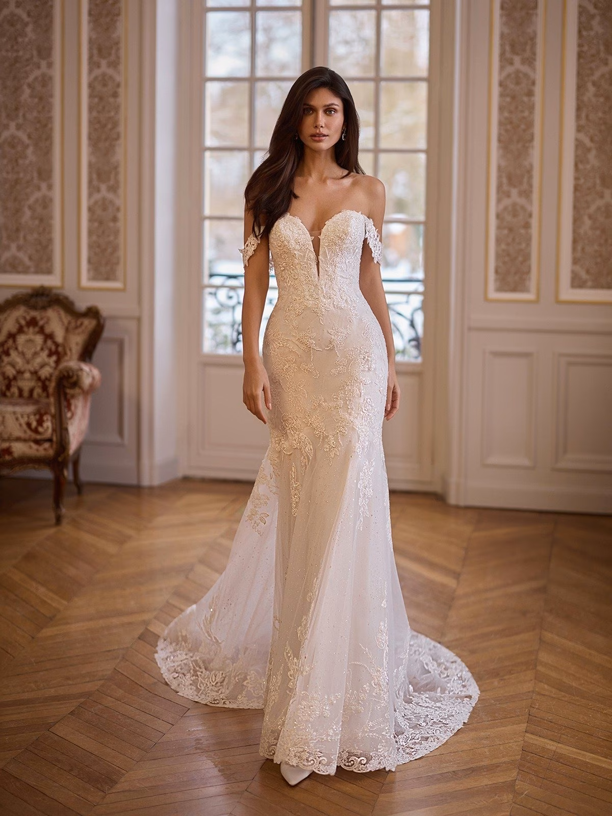 A bride in an elegant, off-the-shoulder sparkly tulle and lace wedding gown stands on polished wooden flooring in a classic room with ornate walls and a large window Moonlight Collection J7011