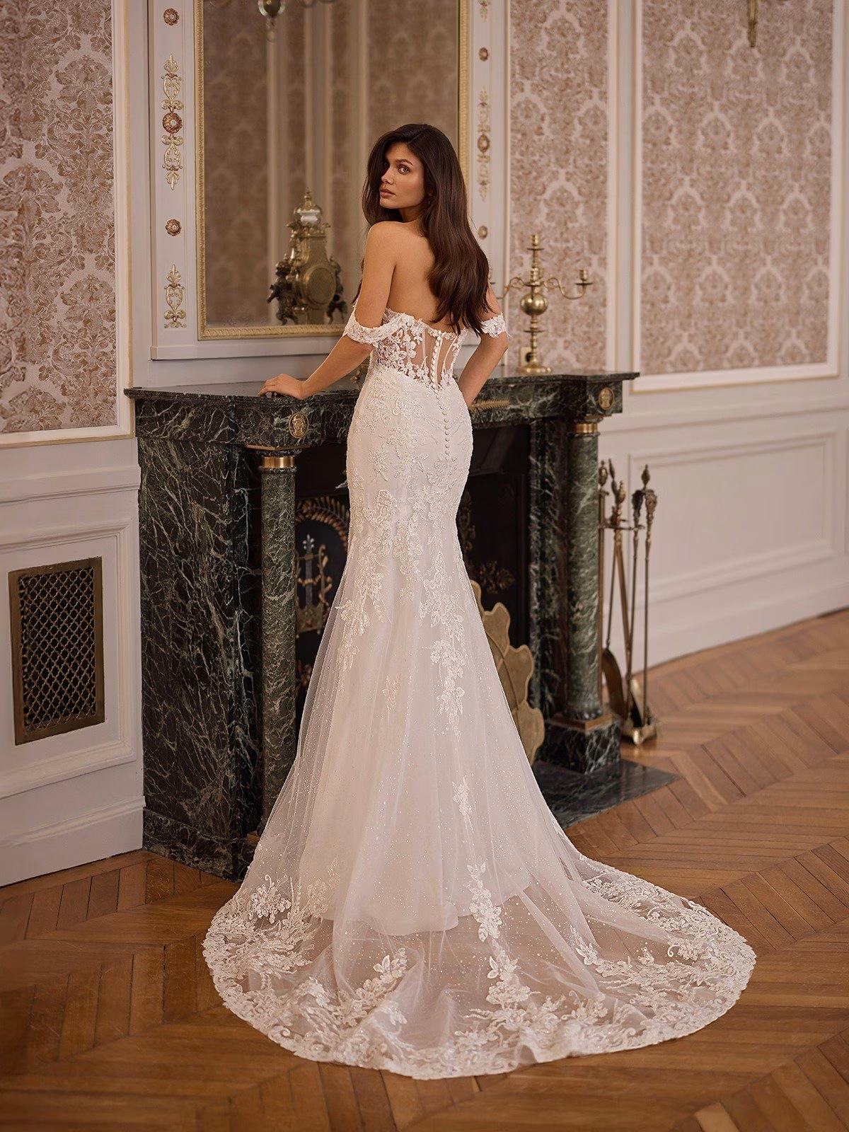A bride in an elegant, lace-trimmed wedding gown with illusion lace train stands by a marble fireplace in a vintage room. Her expression is serene and reflective Moonlight Collection J7011