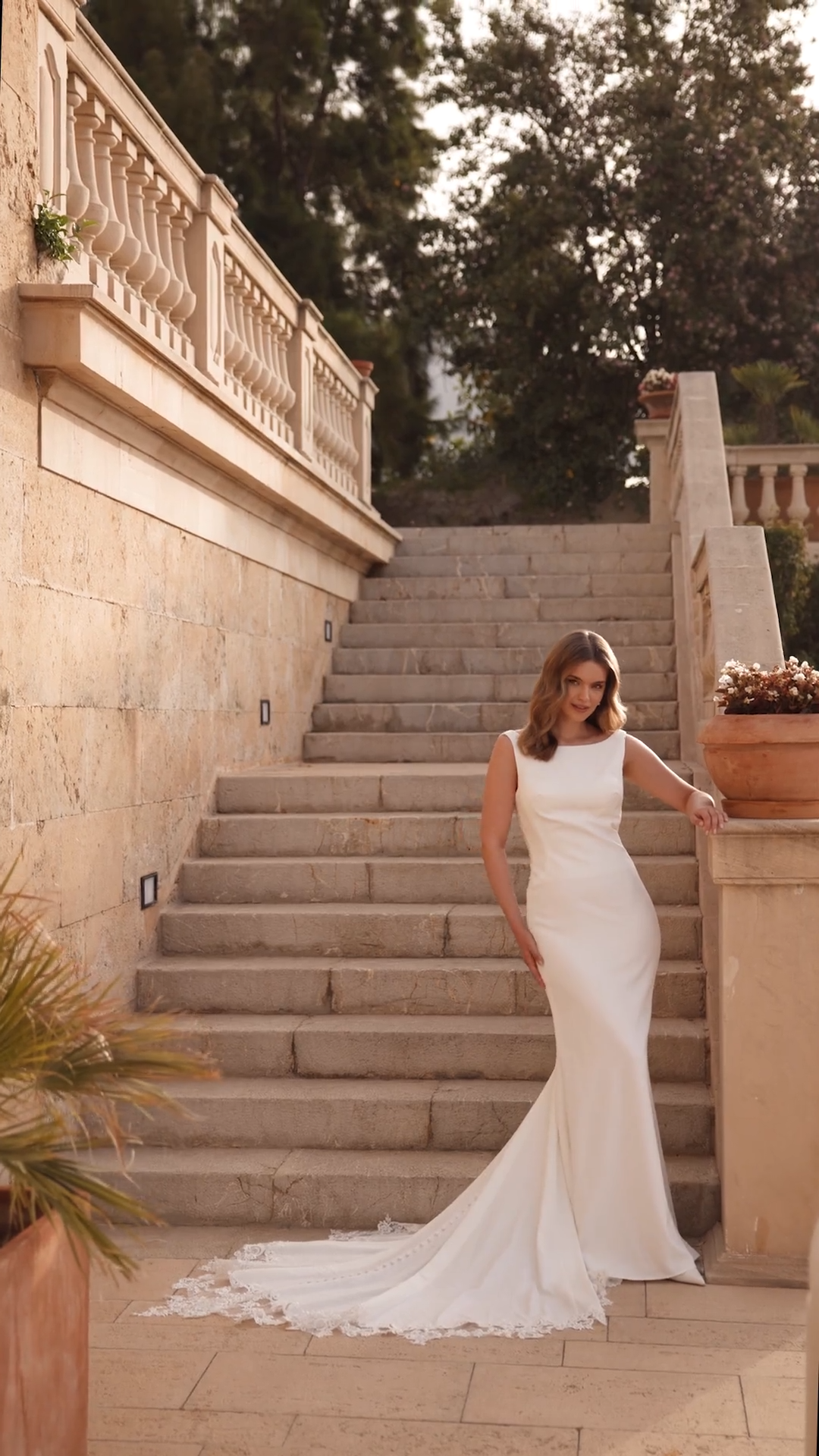 Bride At Bottom Of Staircase Wearing Sleek Column Crepe Wedding Dress With Bateau Neckline