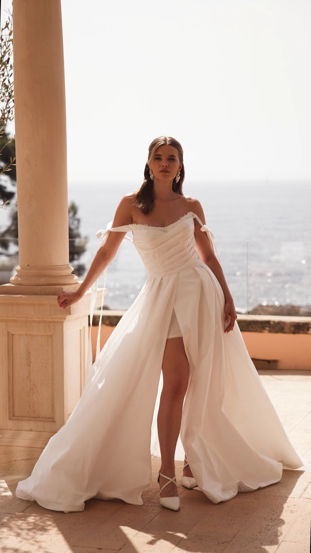 Bride Resting Hand On Column In A Taffeta Wedding Dress With Wrap Front Skirt and Bow Off The Shoulder Sleeves