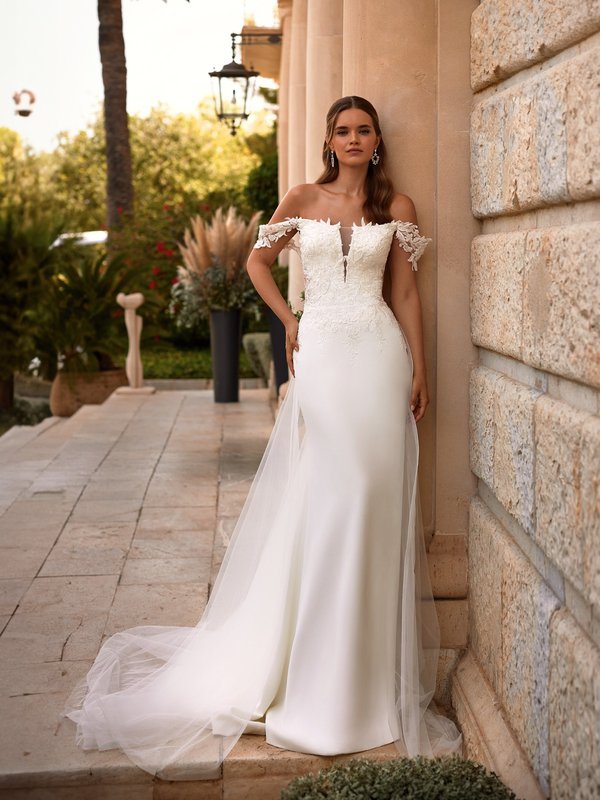 Bride standing next to wall wearing a crepe off the should wedding gown with detachable tulle train