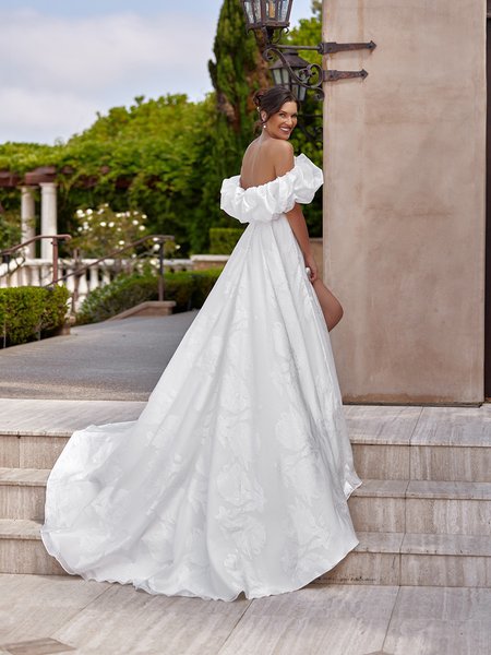 A bride wearing an off-the-shoulder white bridal gown with a dramatic ruffled neckline and a flowing train is standing outdoors on marble steps. She faces away from the camera, turning slightly to smile over her shoulder, highlighting the dress's open back and elegant drape. The fabric features a subtle floral pattern that adds texture and sophistication to the gown. A bride wearing an off-the-shoulder white bridal gown with a dramatic ruffled neckline and a flowing train is standing outdoors on marble steps. She faces away from the camera, turning slightly to smile over her shoulder, highlighting the dress's open back and elegant drape. The fabric features a subtle floral pattern that adds texture and sophistication to the gown.